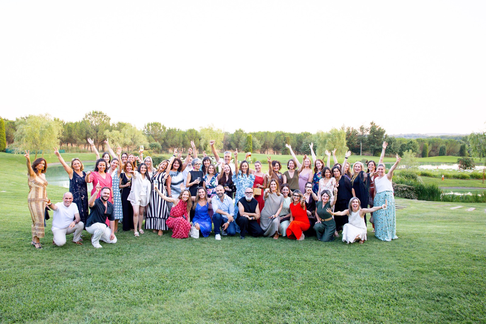 Gran grupo diverso de personas reunidas al aire libre en un campo verde, posando juntos con alegría y levantando los brazos