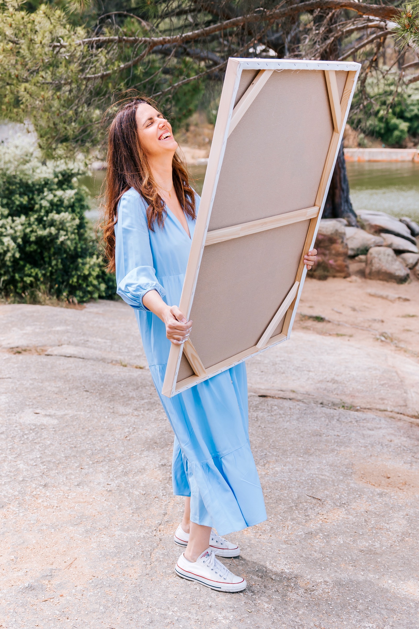 Mujer en vestido azul sosteniendo un lienzo en blanco en un entorno natural cerca de un lago, rodeada de árboles y rocas