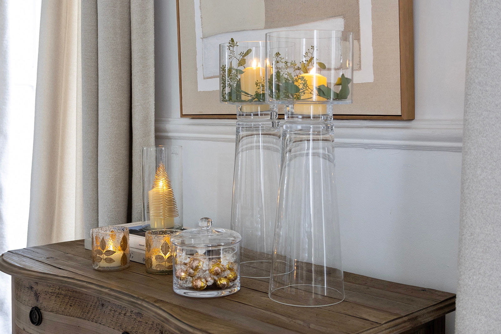 Composición decorativa con velas en recipientes de cristal y jarrones altos transparentes sobre una cómoda de madera rústica. Las velas encendidas crean un ambiente cálido y acogedor con detalles dorados y vegetales.