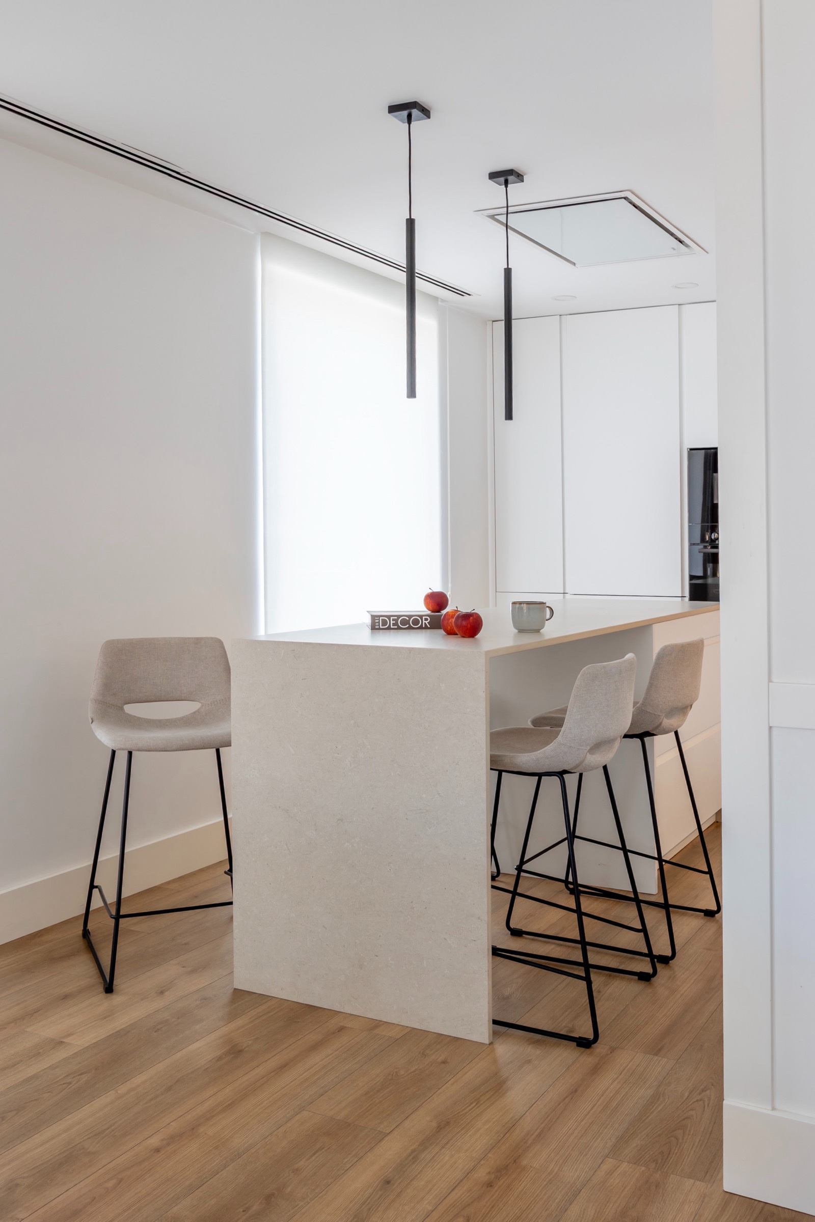Espacio de cocina minimalista con barra blanca, tres bancos altos beige, manzanas rojas y lámpara colgante delgada
