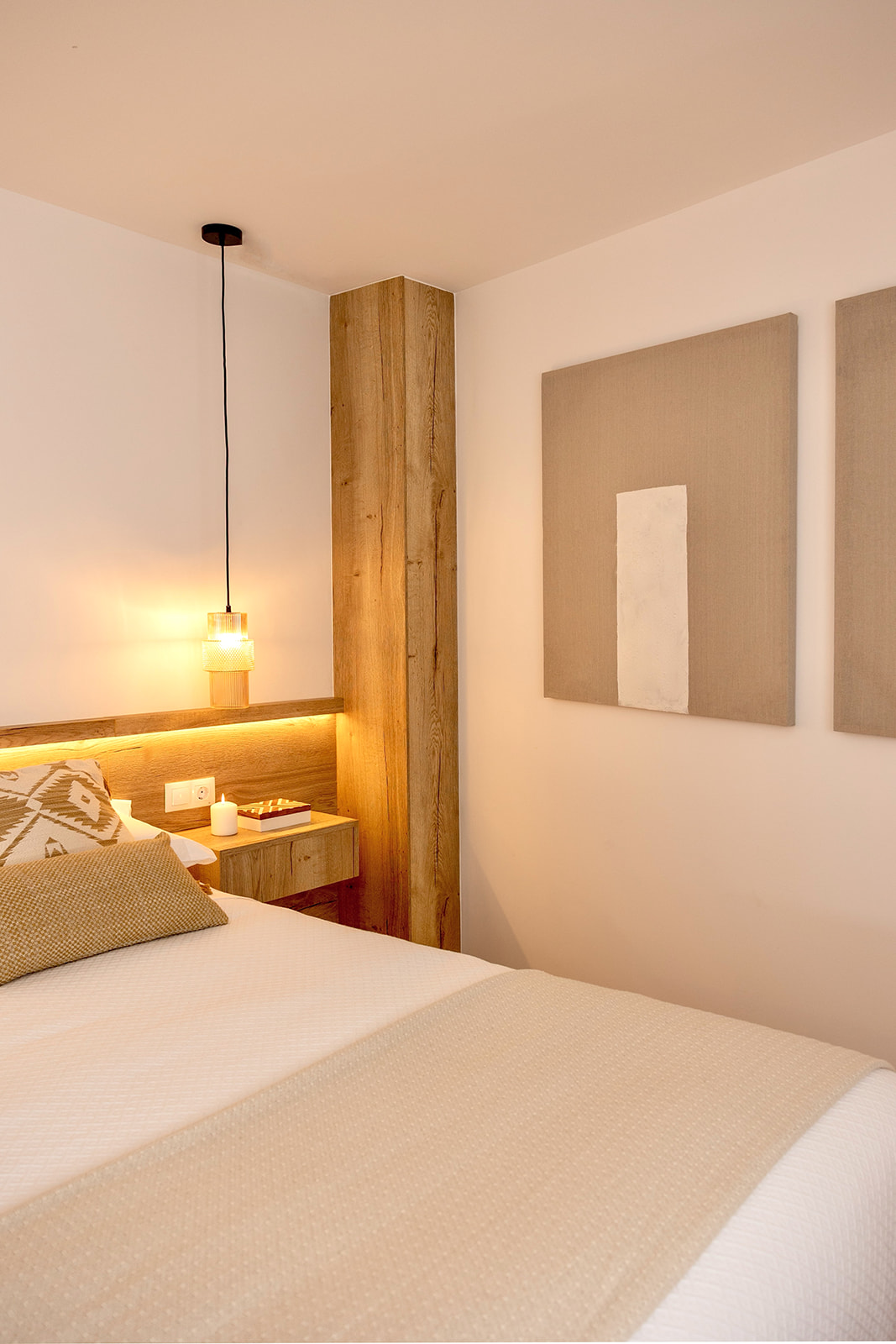 Dormitorio moderno con tonos cálidos, cama con cabecera de madera iluminada, lámpara colgante, columna de madera y cuadros minimalistas en la pared. Ambiente acogedor y elegante.