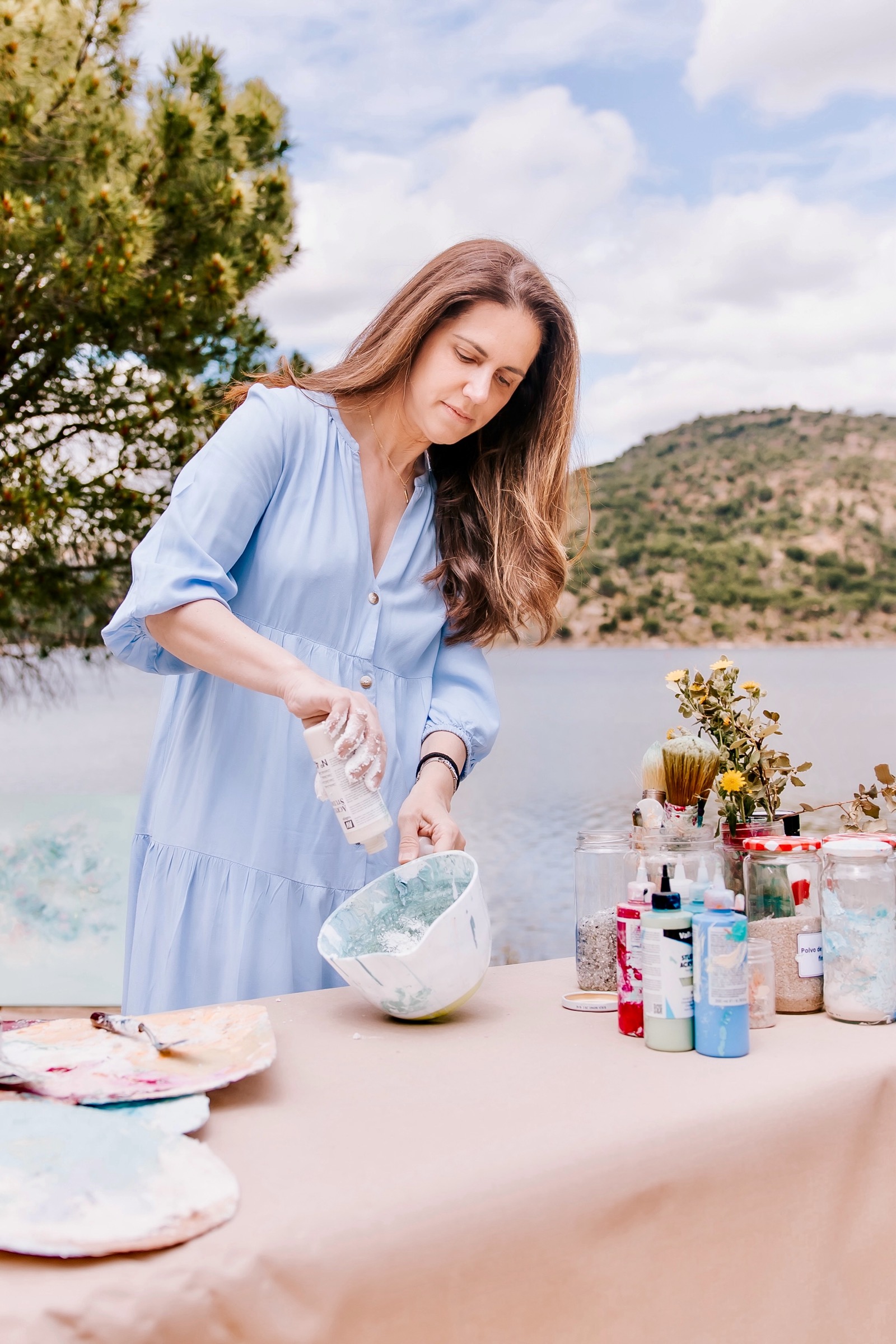 Mujer en vestido azul trabajando en un proyecto de arte junto a un lago, con montañas en el fondo. Tiene materiales artísticos como pinturas y pinceles sobre una mesa frente a ella.