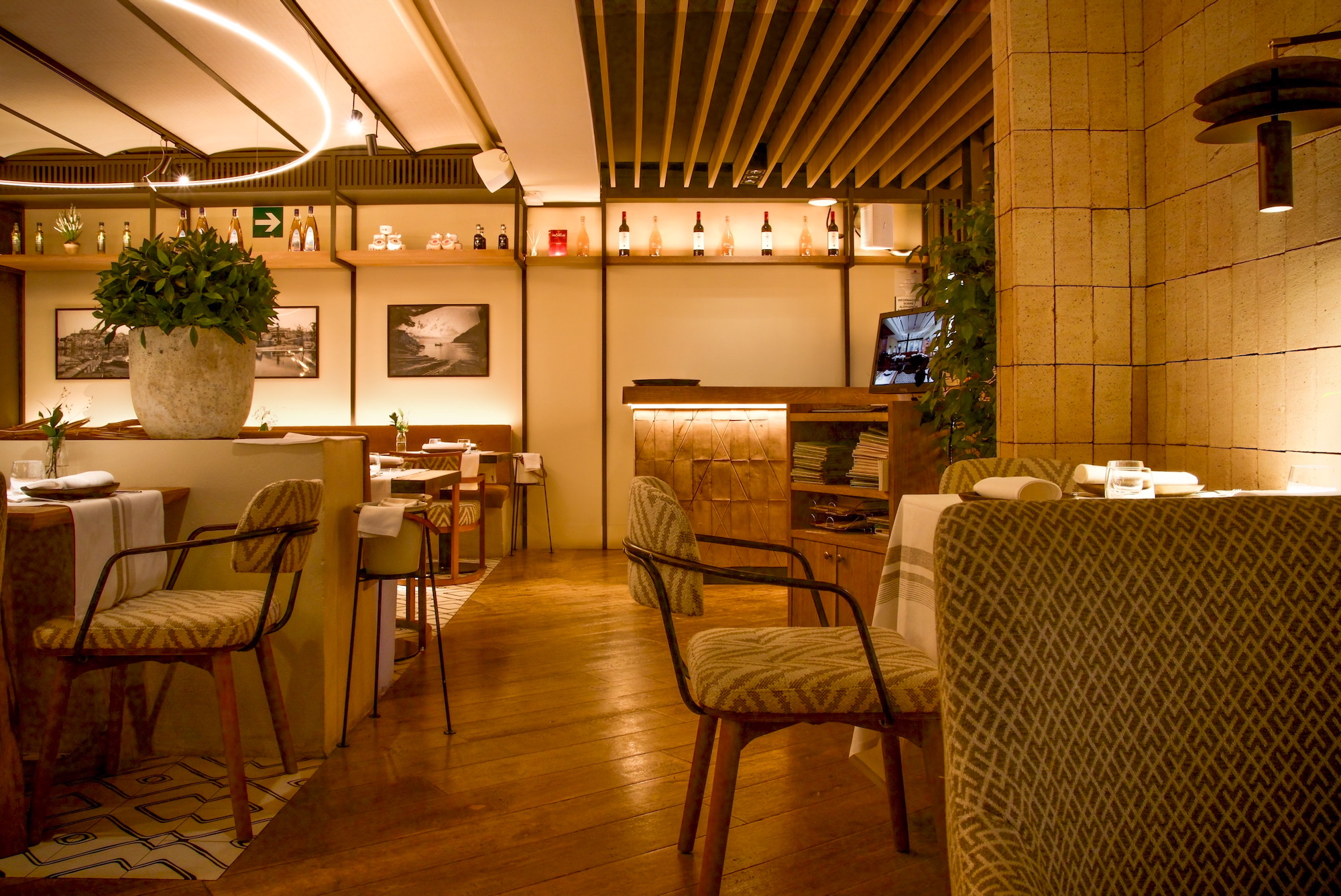 Interior de restaurante elegante con iluminación cálida, sillas tapizadas en verde, suelos de madera, plantas decorativas y fotografías en blanco y negro en las paredes. Ambiente acogedor con detalles modernos y elementos de diseño.