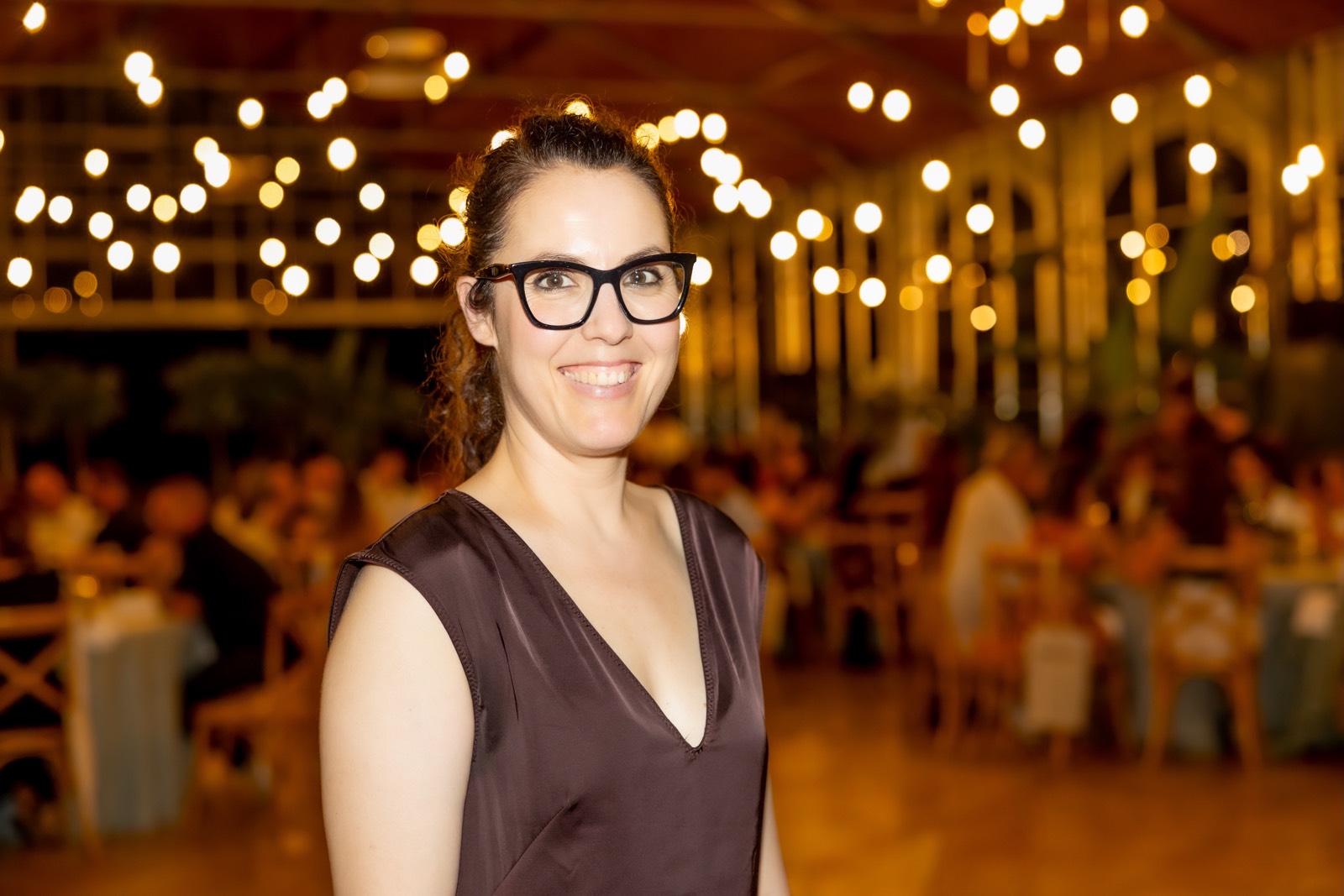 Mujer con gafas sonriendo, vistiendo vestido marrón oscuro, de pie en un espacio con luces cálidas y decorativas desenfocadas