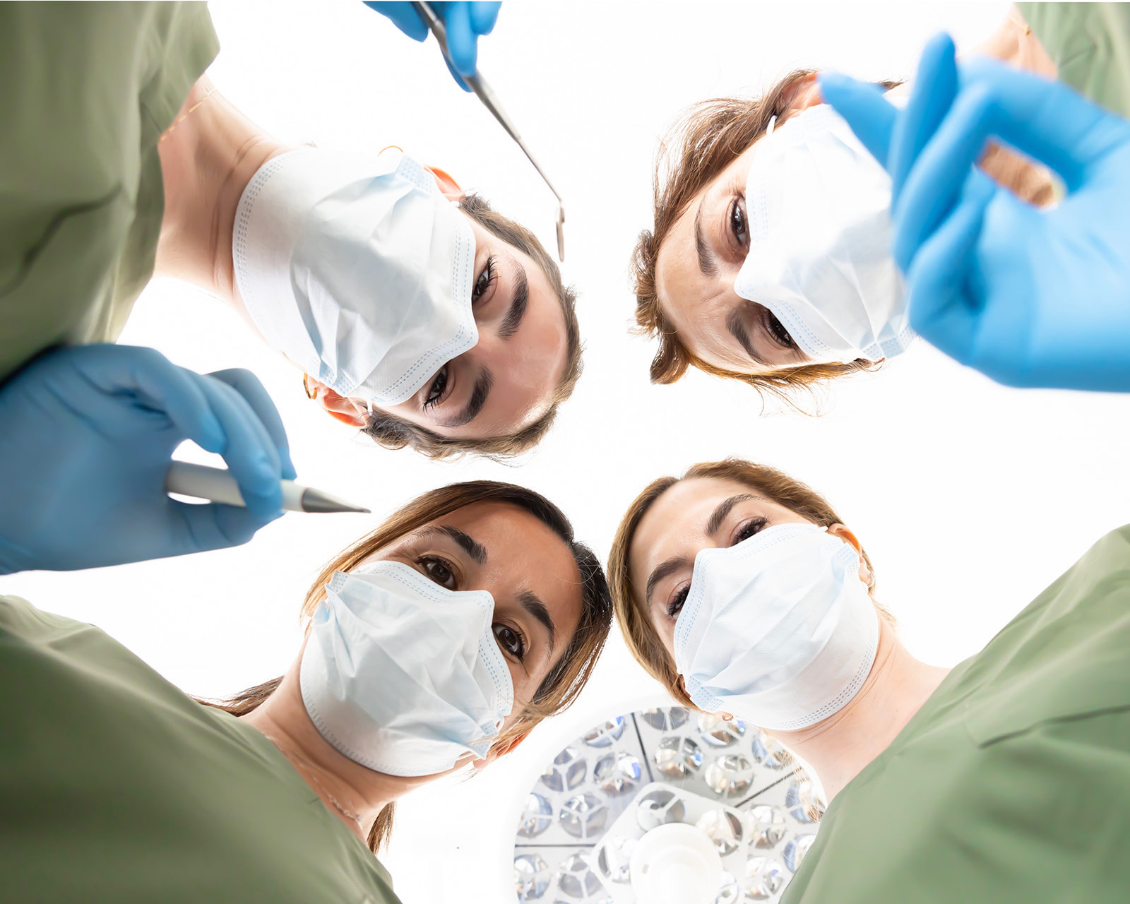 Vista desde abajo de cuatro profesionales médicos con mascarillas y guantes quirúrgicos mirando hacia la cámara. Están rodeando una lámpara quirúrgica, sugiriendo que están en un quirófano preparados para una operación.