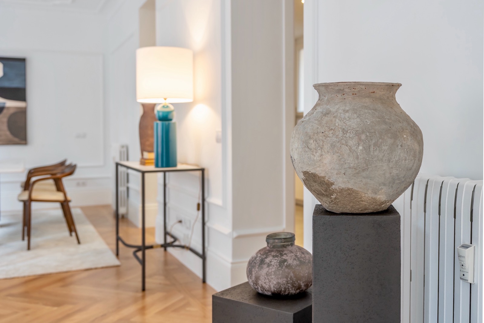 Interior de sala de estar moderna con decoración minimalista, incluyendo jarrones antiguos sobre pedestales, una mesa auxiliar con lámpara de diseño, y sillas de madera al fondo. Paredes blancas y suelo de madera.