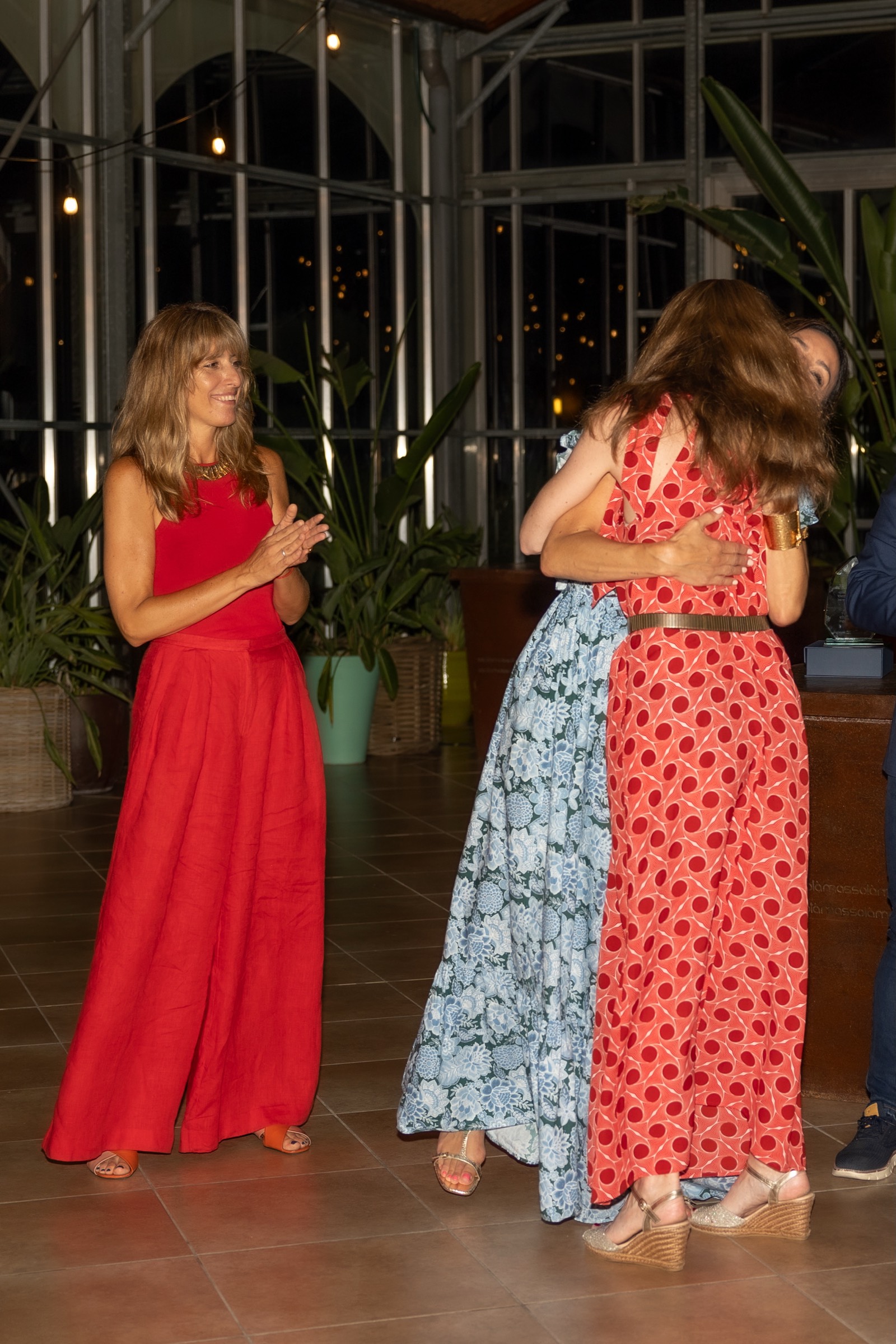 Dos mujeres abrazándose en un evento nocturno con vestidos coloridos, una de rojo y otra de rojo con lunares, en un ambiente con plantas y luces
