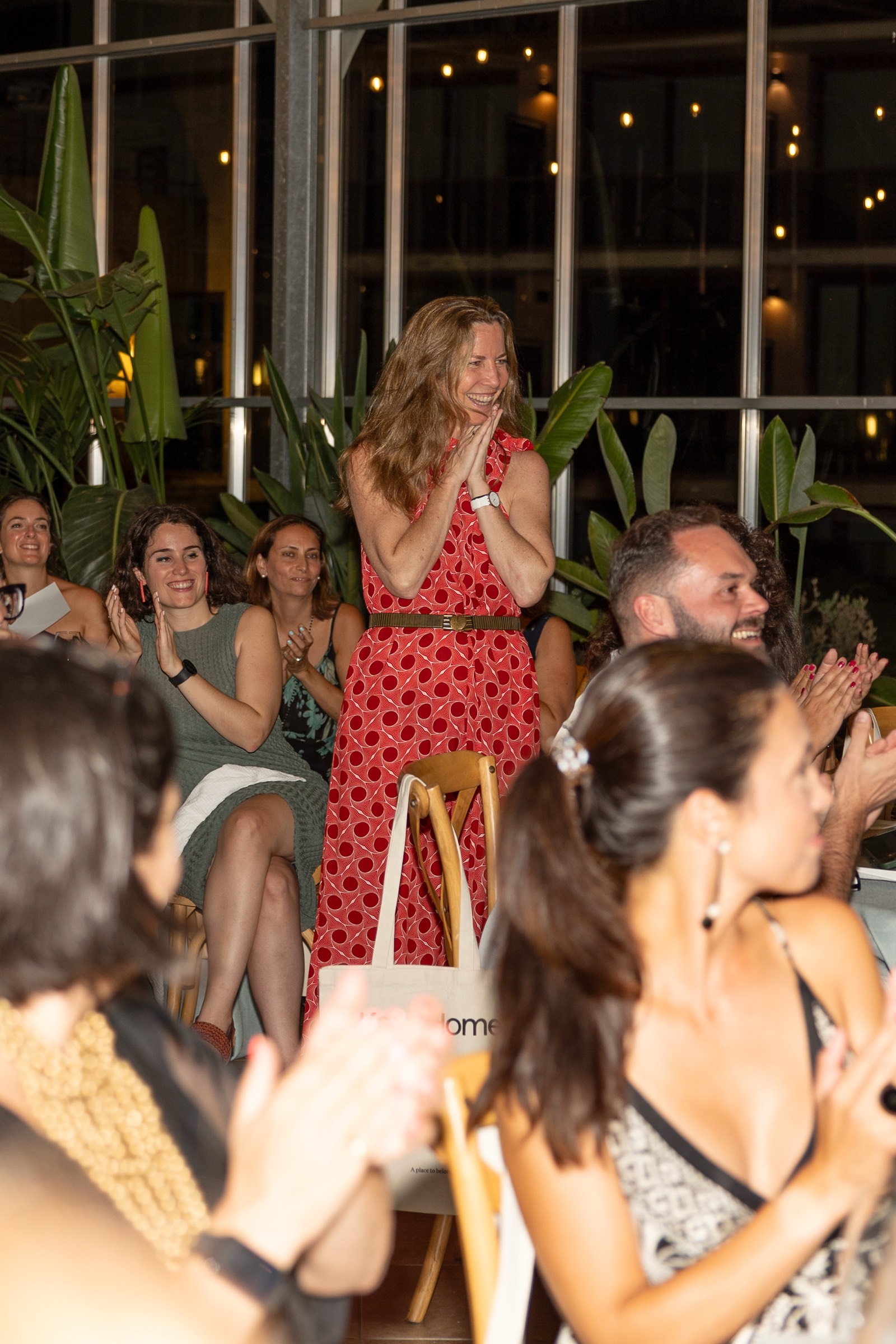Grupo de mujeres en un evento social, una mujer con vestido rojo está de pie aplaudiendo, rodeada de plantas y luces cálidas