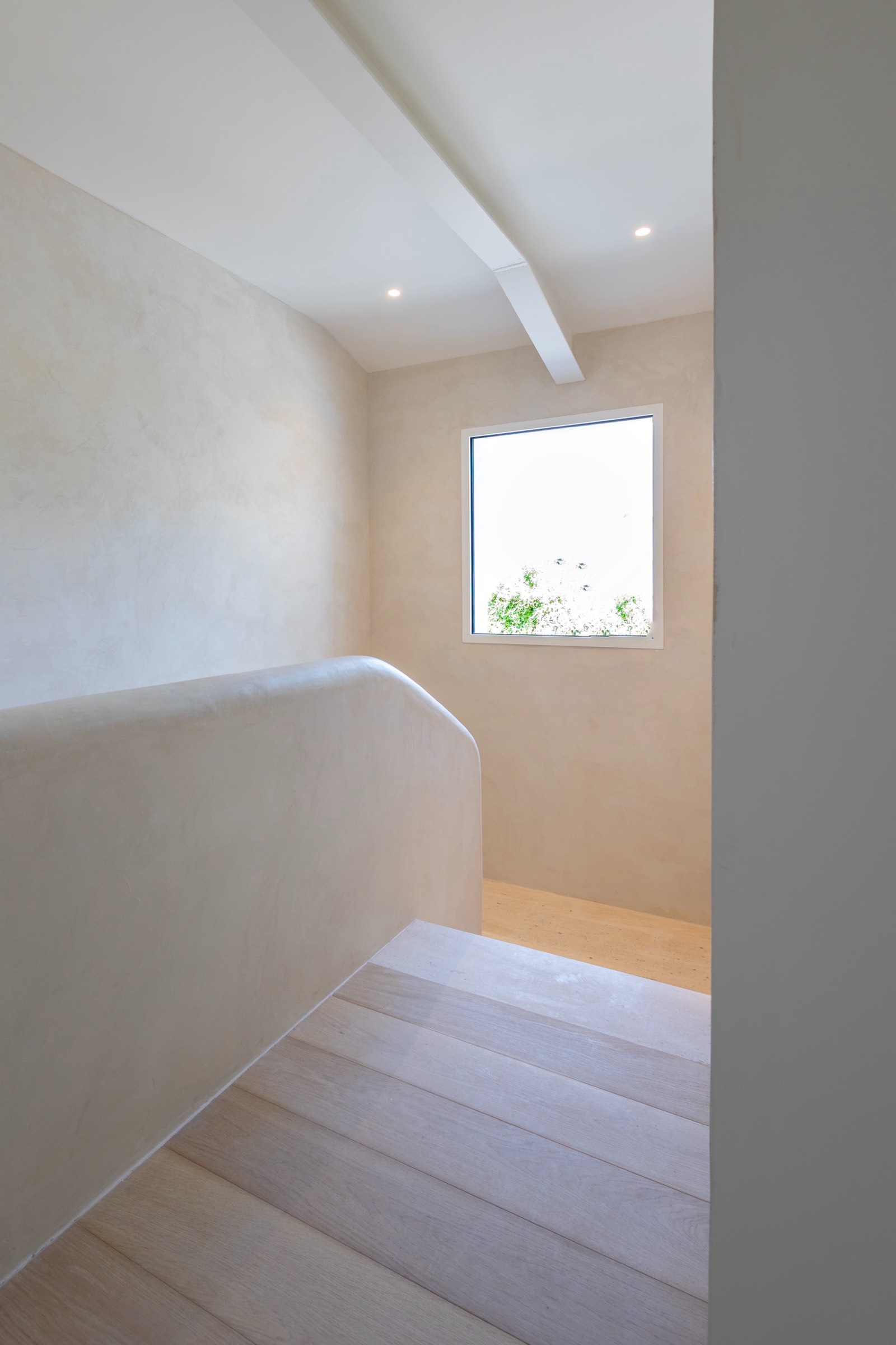 Interior de pasillo luminoso con piso de madera clara, paredes beige suaves, recorte de ventana con vista a follaje verde y luz natural