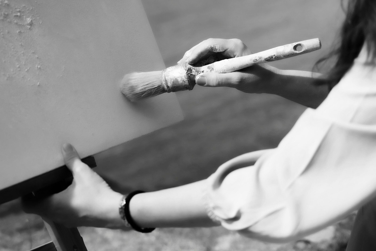 Imagen en blanco y negro de una persona sosteniendo una brocha mientras pinta una pared. Se enfoca en las manos del pintor y la brocha en movimiento, mostrando el proceso de pintura en acción.