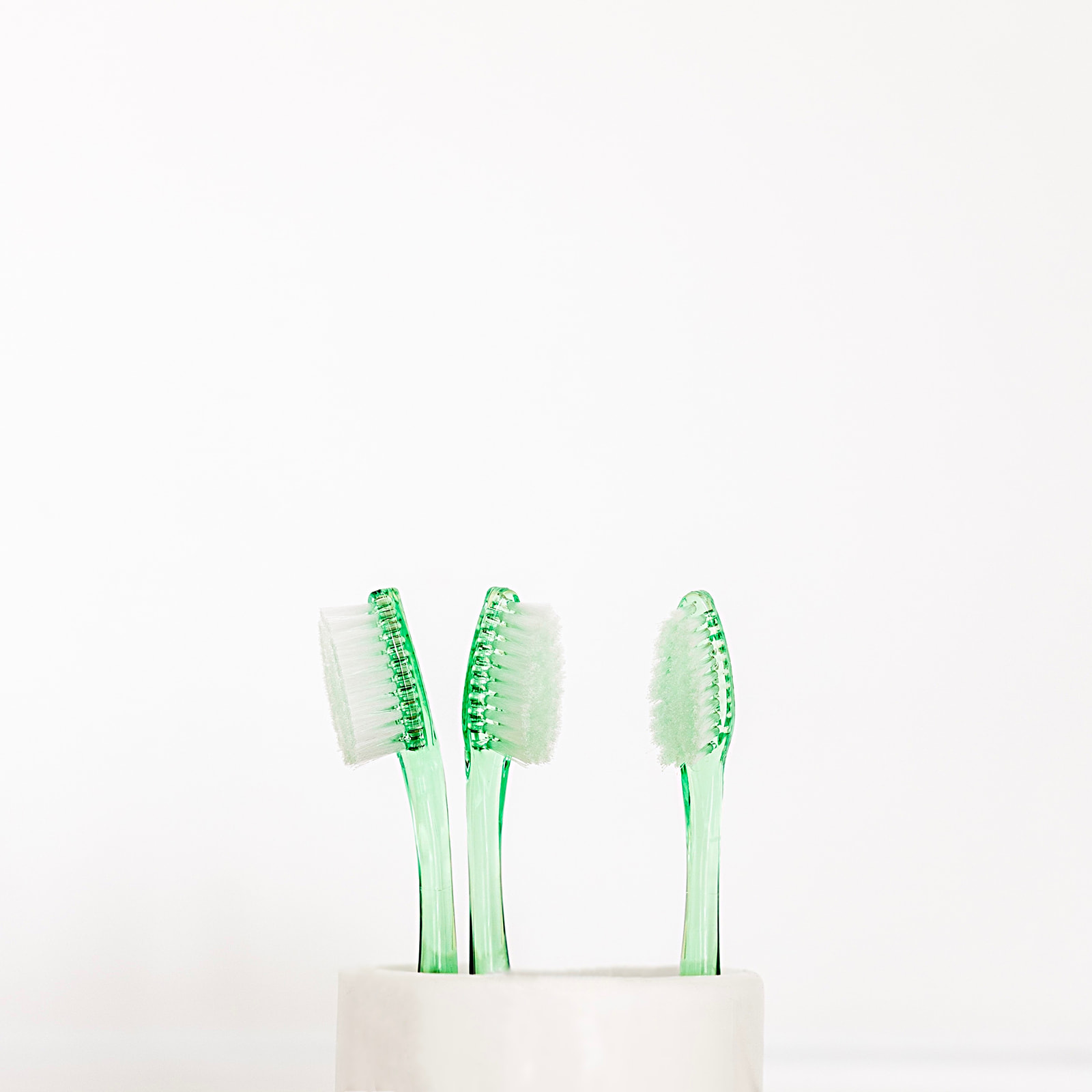 Tres cepillos de dientes transparentes de color verde claro colocados en un soporte blanco sobre un fondo blanco. Los cepillos tienen cerdas blancas y muestran diferentes ángulos.