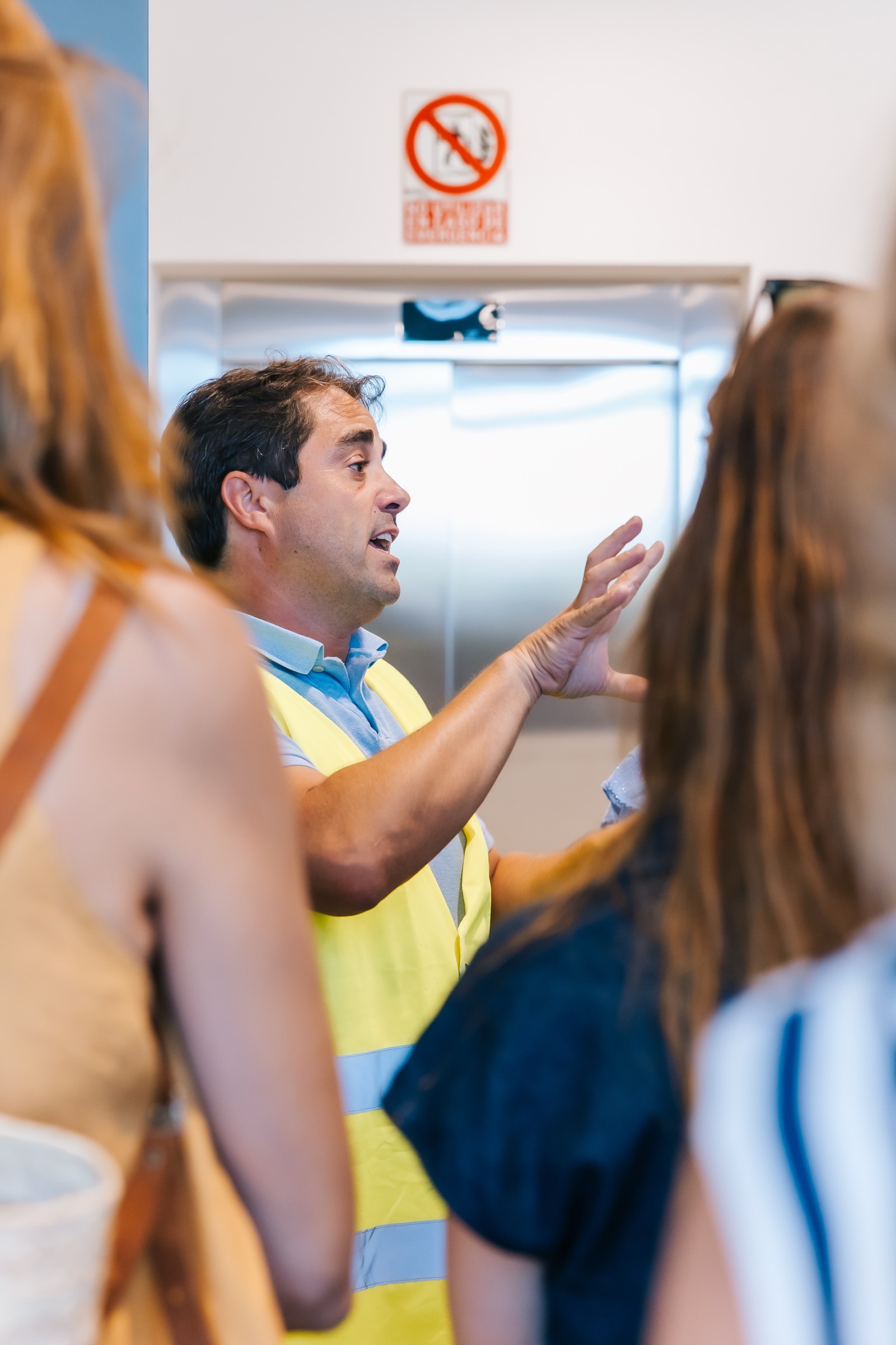 Trabajador con chaleco de seguridad amarillo explica algo gesticulando, con una señal de prohibición en el fondo