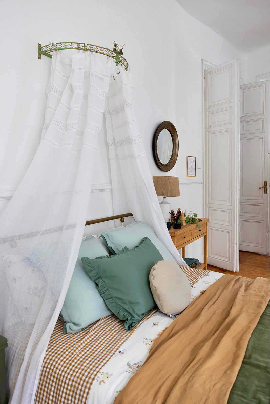 Dormitorio con estilo romántico decorado con dosel blanco sobre la cama, almohadones verde menta, ropa de cama a cuadros beige y detalles en madera natural. Paredes blancas y puertas clásicas.