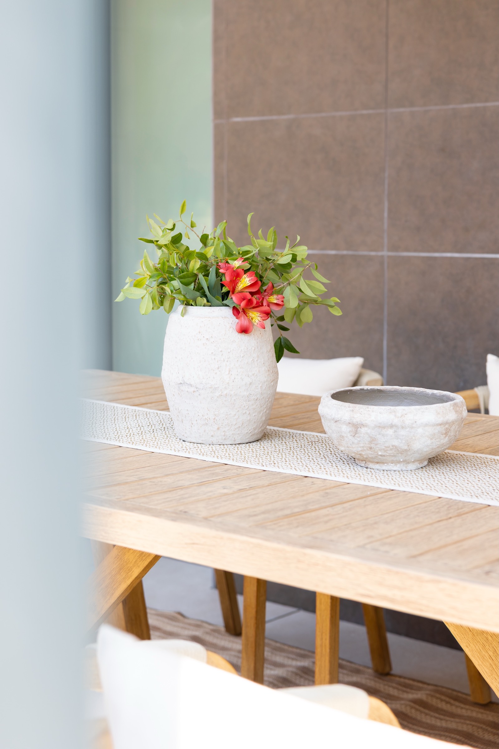 Mesa de comedor de madera con un jarrón blanco conteniendo flores verdes y rojas, y un cuenco decorativo. La mesa está cubierta por un camino de mesa blanco y se encuentra en una habitación con paredes de azulejos marrones.