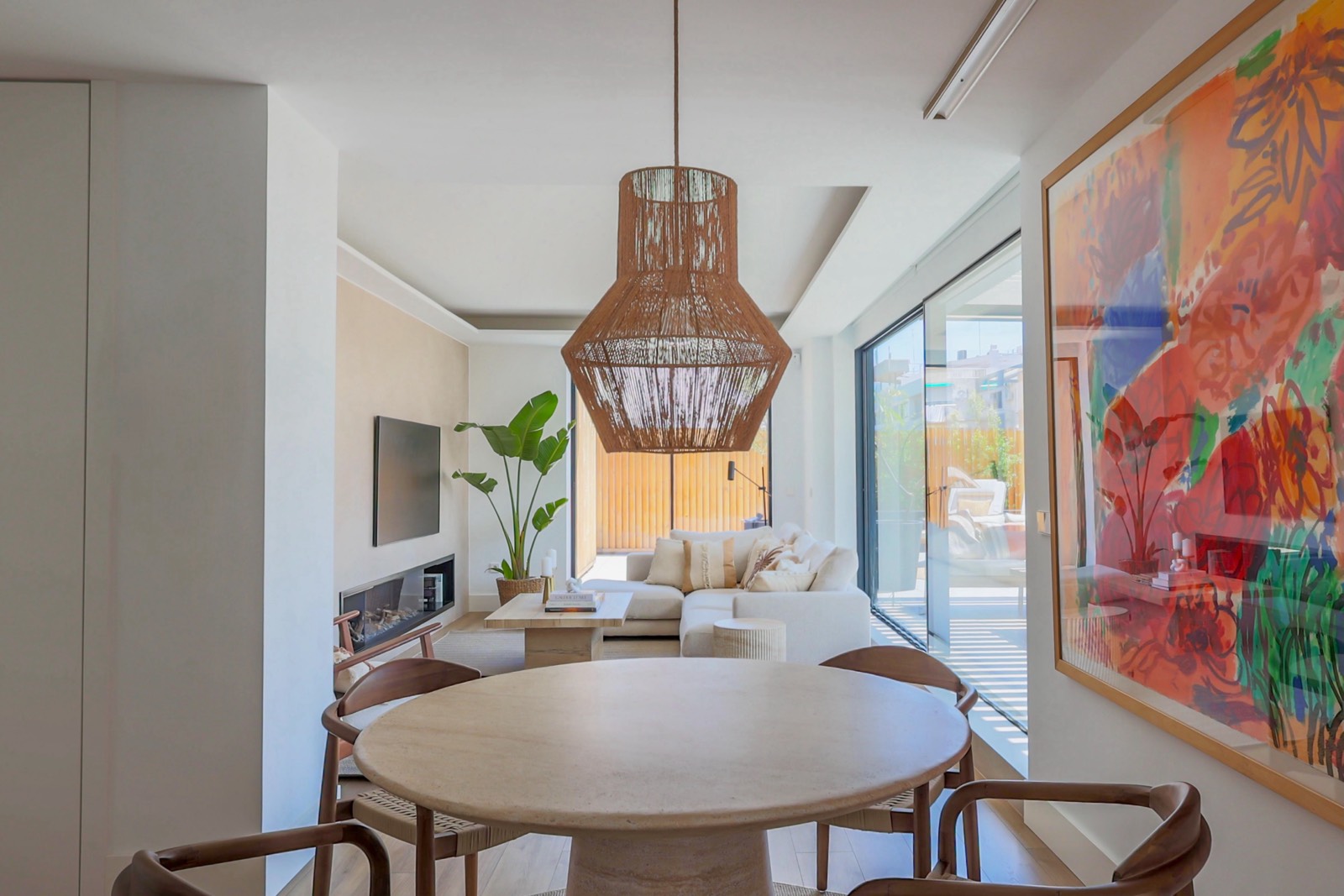Sala de estar y comedor modernos con gran ventanal, lámpara colgante de ratán, sofá blanco, mesa redonda de comedor y cuadro colorido en la pared. El espacio es luminoso y tiene un estilo contemporáneo con toques naturales.