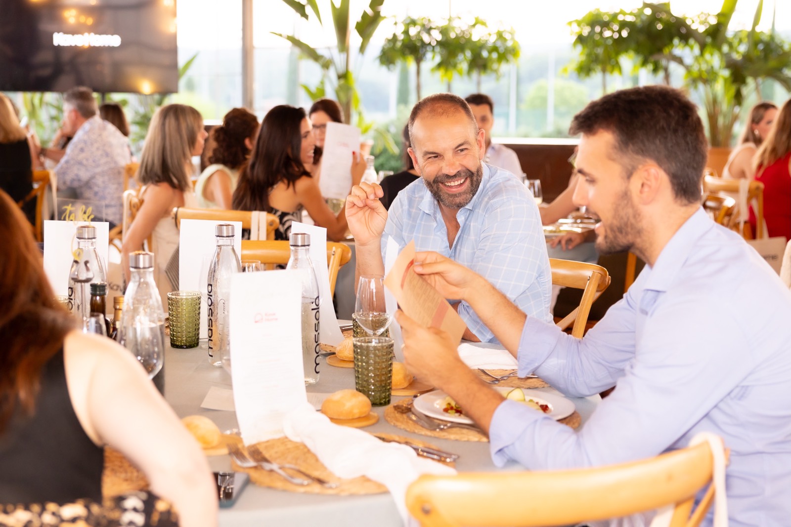 Grupo de personas cenando juntas en un restaurante luminoso con plantas, conversando animadamente durante un almuerzo de trabajo