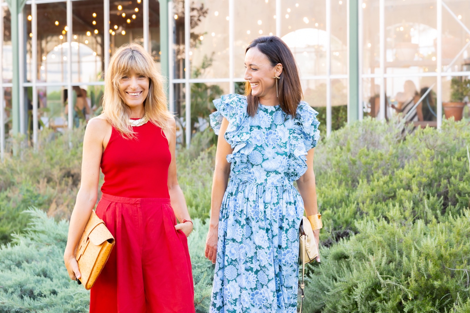 Dos mujeres sonrientes vestidas elegantemente, una con vestido rojo y otra con vestido azul floral, de pie en un invernadero con plantas verdes