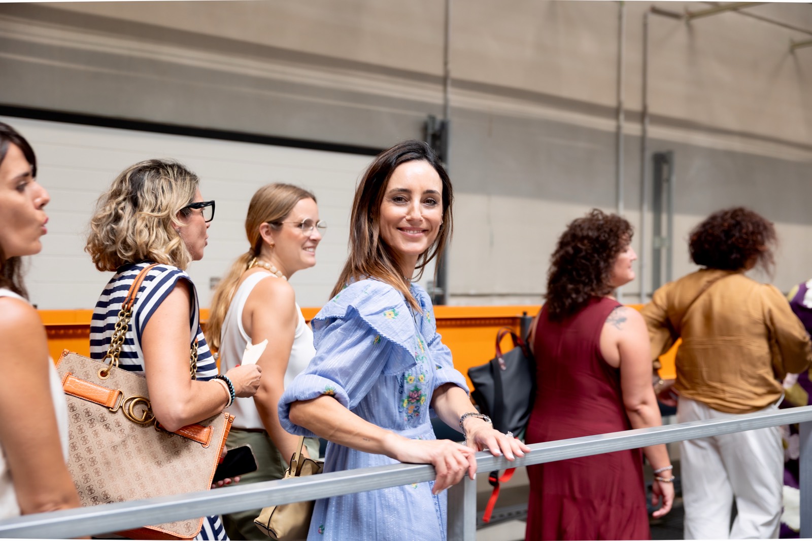 Grupo de mujeres en fila, de pie junto a una barandilla, vistiendo diferentes estilos de ropa, algunas con bolsos y gafas