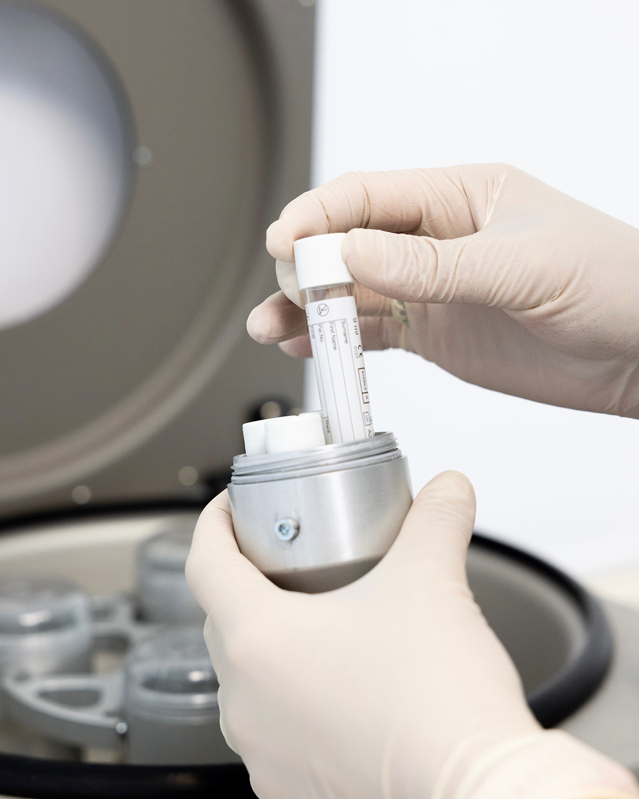 Manos con guantes de látex manipulando un tubo de ensayo y un contenedor metálico en un entorno de laboratorio. Se ve parte de un equipo de laboratorio en el fondo.