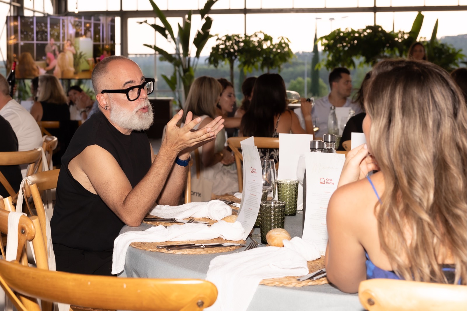 Escena de almuerzo en un restaurante con un hombre mayor con gafas y barba blanca conversando animadamente en una mesa con otras personas