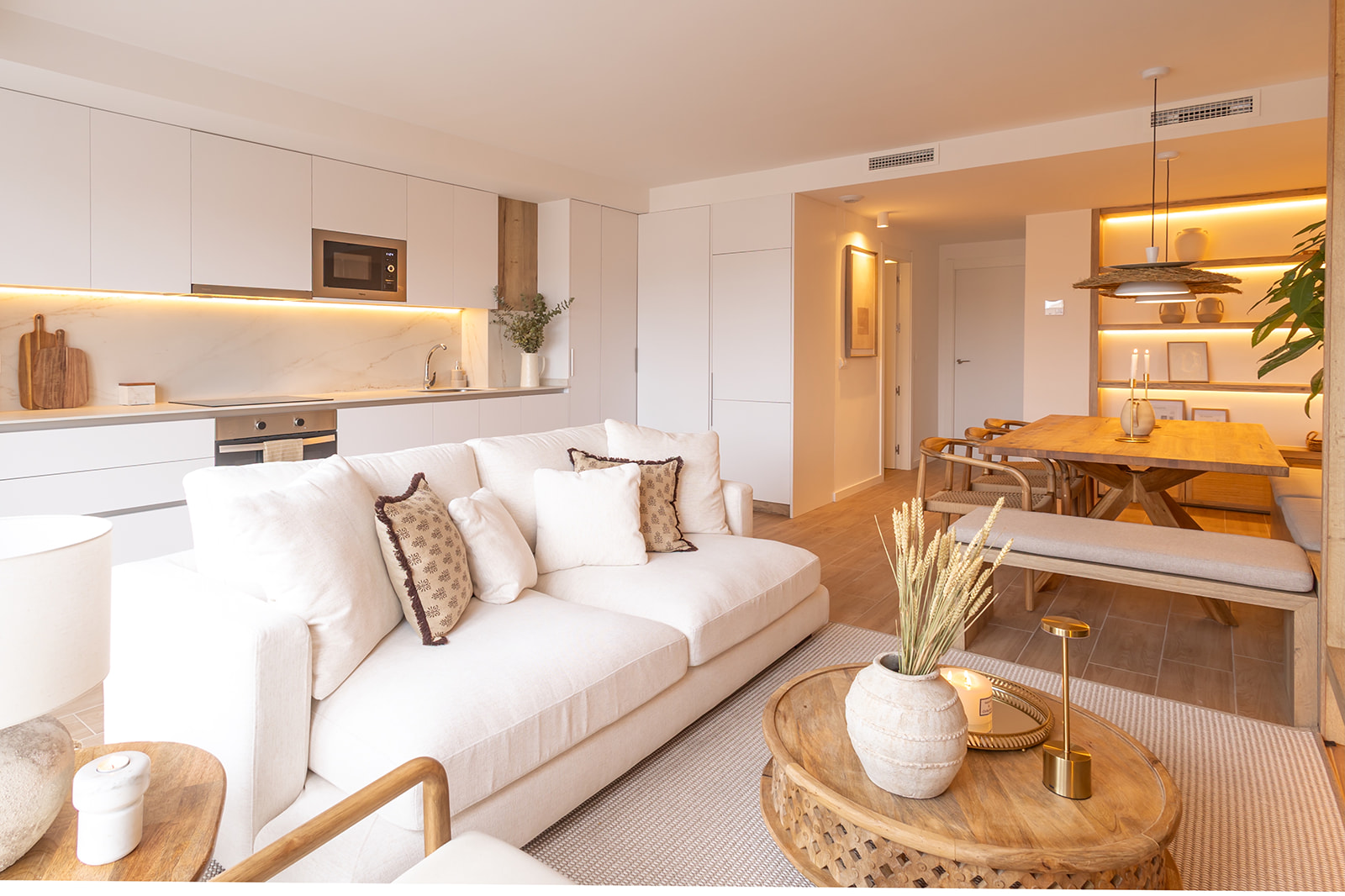 Apartamento moderno de planta abierta con cocina, sala de estar y comedor en tonos claros. Decoración minimalista con acentos de madera y iluminación cálida. Sofá blanco, mesa de centro rústica y comedor de madera.
