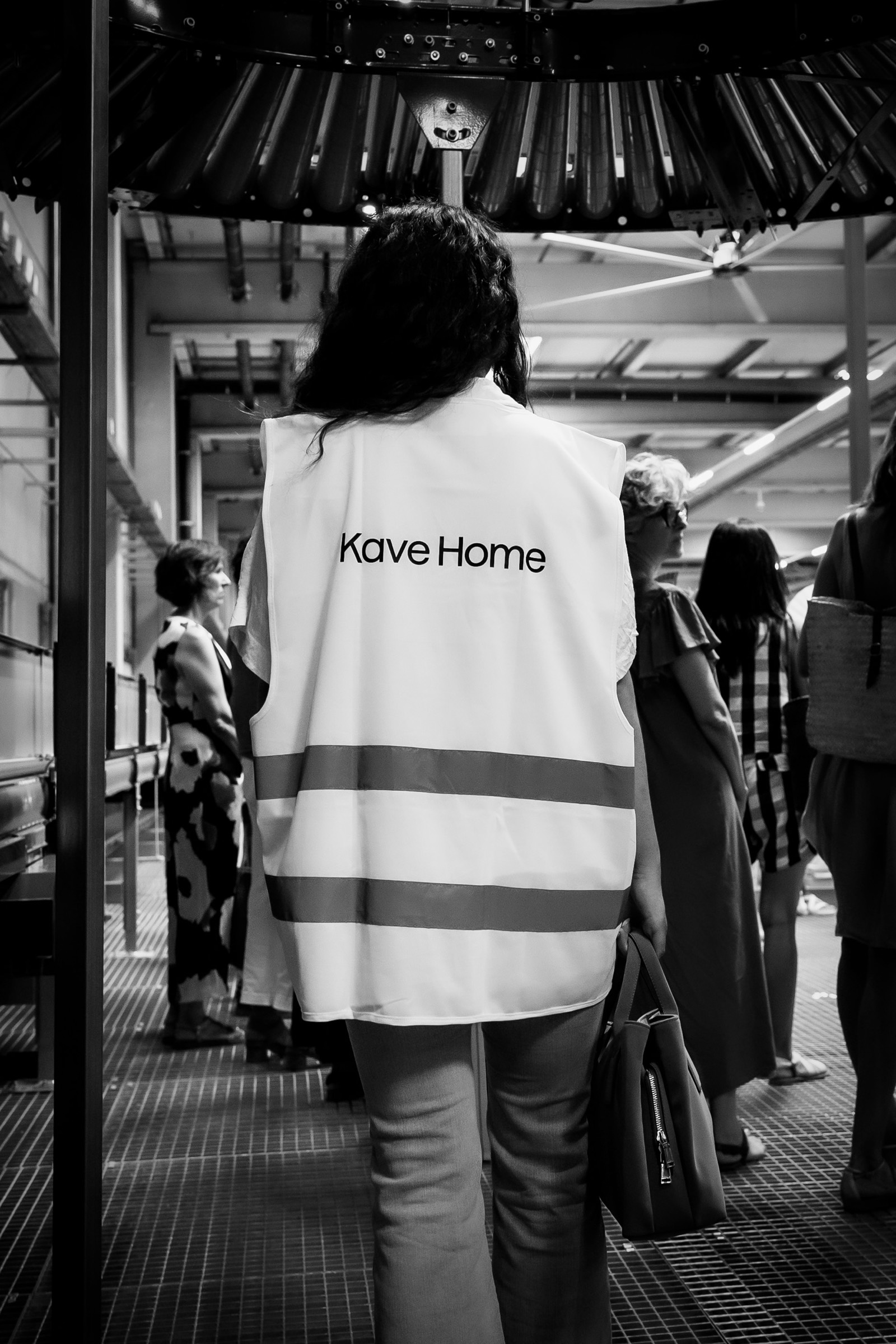 Vista trasera de un empleado con chaleco reflectante blanco de Kave Home en un entorno industrial, rodeado de otras personas