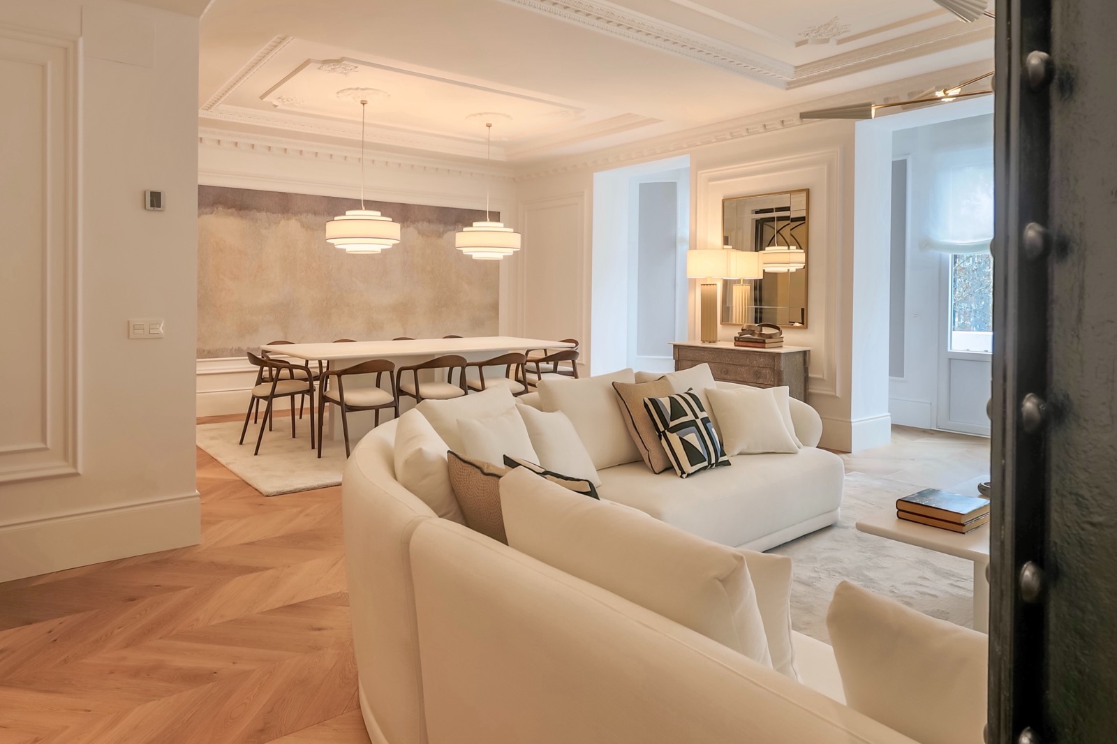 Espaciosa sala de estar y comedor con decoración elegante y moderna. Sofá curvo blanco, mesa de comedor grande, iluminación colgante y suelos de parquet. Techos altos con molduras ornamentales y paredes blancas.