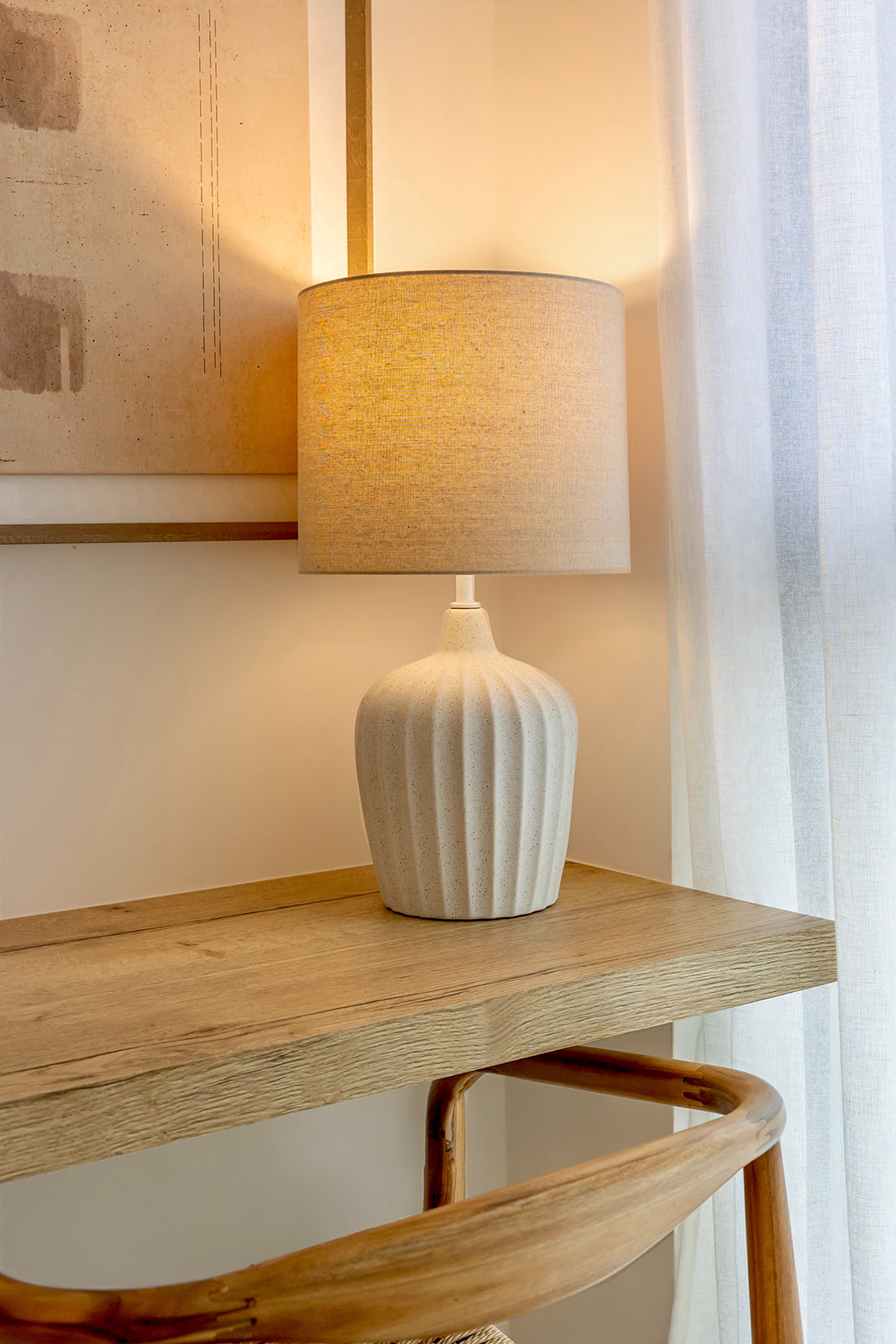 Lámpara de mesa con base de cerámica blanca estriada y pantalla de tela beige sobre un escritorio de madera clara junto a una ventana con cortinas translúcidas. La luz cálida crea una atmósfera acogedora en la habitación.