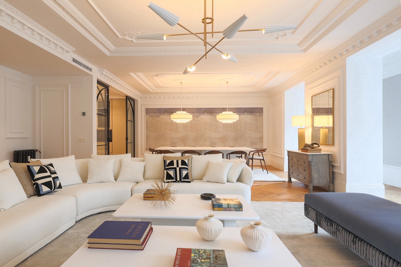 Sala de estar espaciosa y elegante con decoración moderna. Sofá blanco curvo, mesas de centro blancas, lámpara de techo geométrica y comedor al fondo. Paredes blancas con molduras ornamentadas y suelo de madera.