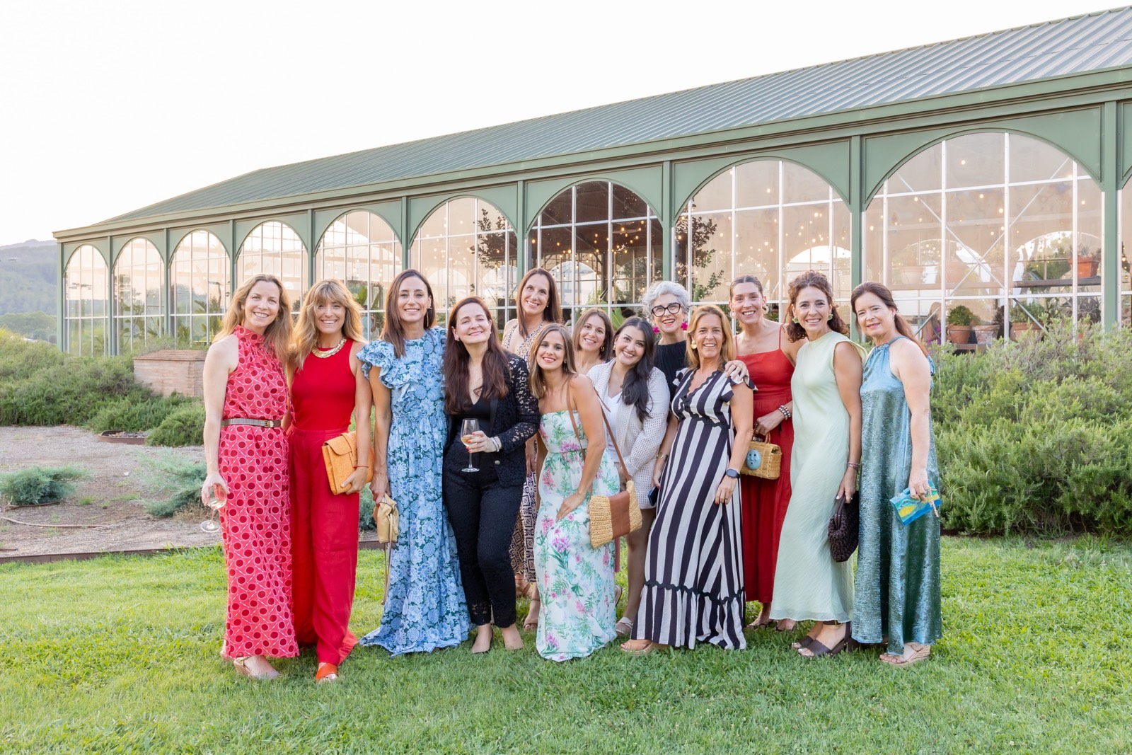 Grupo de mujeres vestidas elegantemente posando juntas frente a un edificio de cristal con jardines verdes