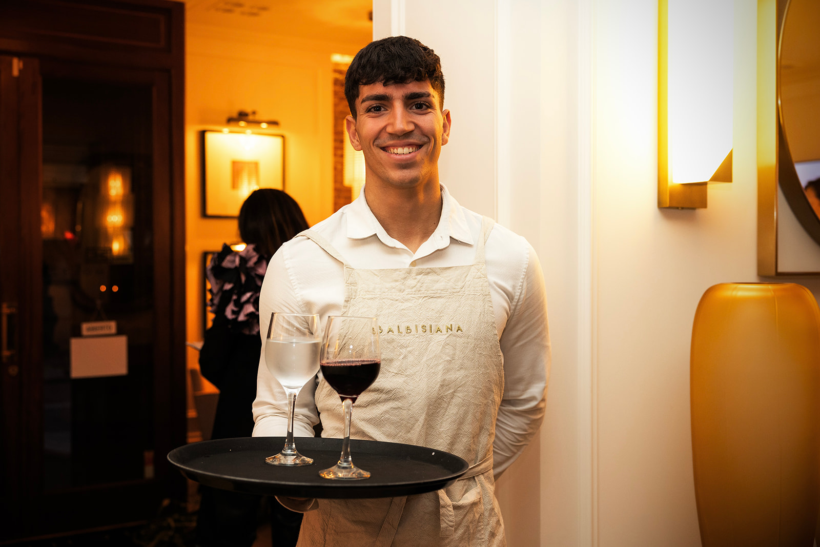 Camarero sonriente en un restaurante elegante, vistiendo camisa blanca y delantal con el nombre 'Dalbisiana', sosteniendo una bandeja con una copa de vino tinto y una de vino blanco