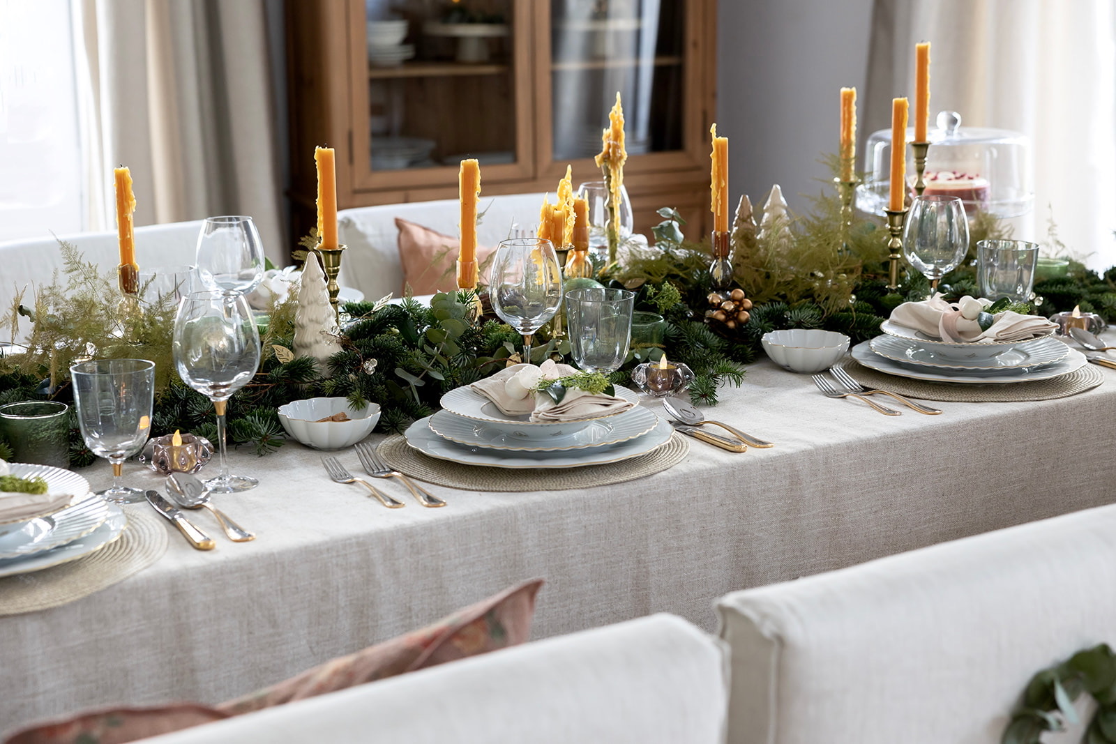 Mesa navideña decorada con un centro de mesa de ramas de pino, velas doradas altas, cristalería elegante, vajilla blanca y gris sobre mantel de lino beige, con detalles decorativos festivos y una disposición formal