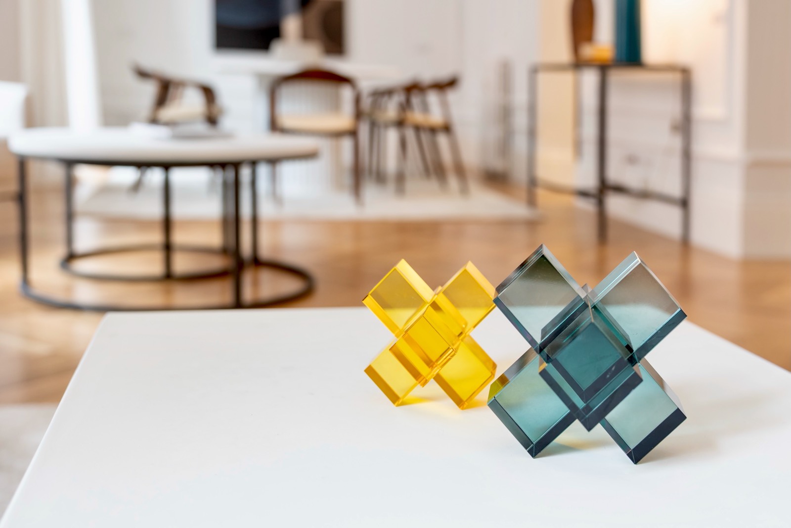 Dos esculturas geométricas, una amarilla y otra verde azulada, sobre una mesa blanca en una sala de exposición con muebles modernos al fondo
