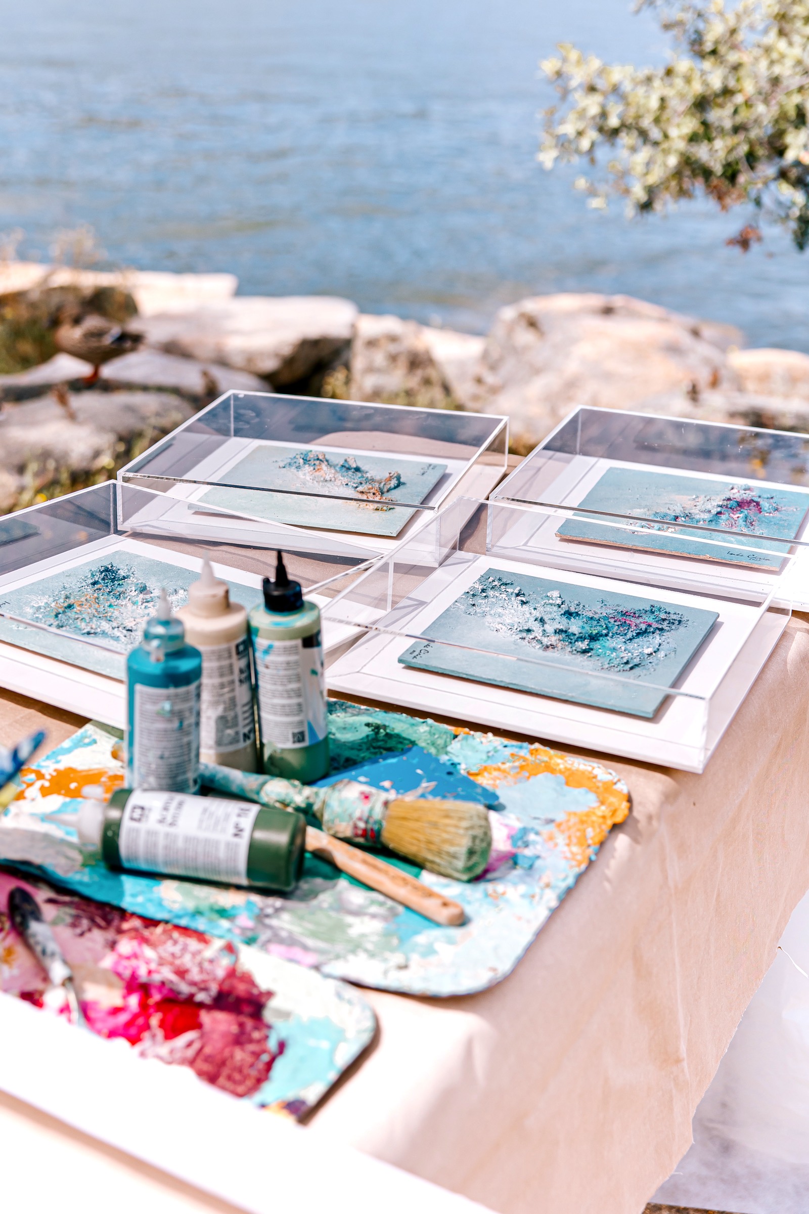 Mesa al aire libre con materiales de pintura y lienzos cerca del mar. Se ven pinturas en proceso, pinceles, tubos de pintura y paletas coloridas. El fondo muestra rocas y agua azul, creando un ambiente artístico y sereno.