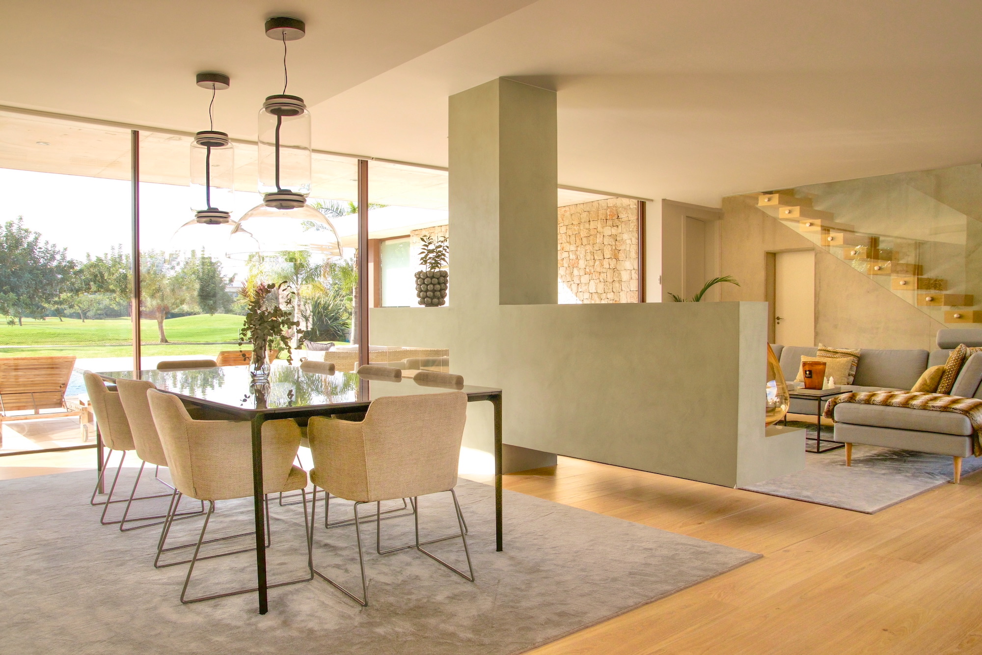 Comedor moderno y elegante con mesa negra y sillas beige, integrado en espacio abierto con sala de estar. Amplios ventanales que permiten entrada de luz natural y vista al jardín. Diseño minimalista con lámparas colgantes decorativas.
