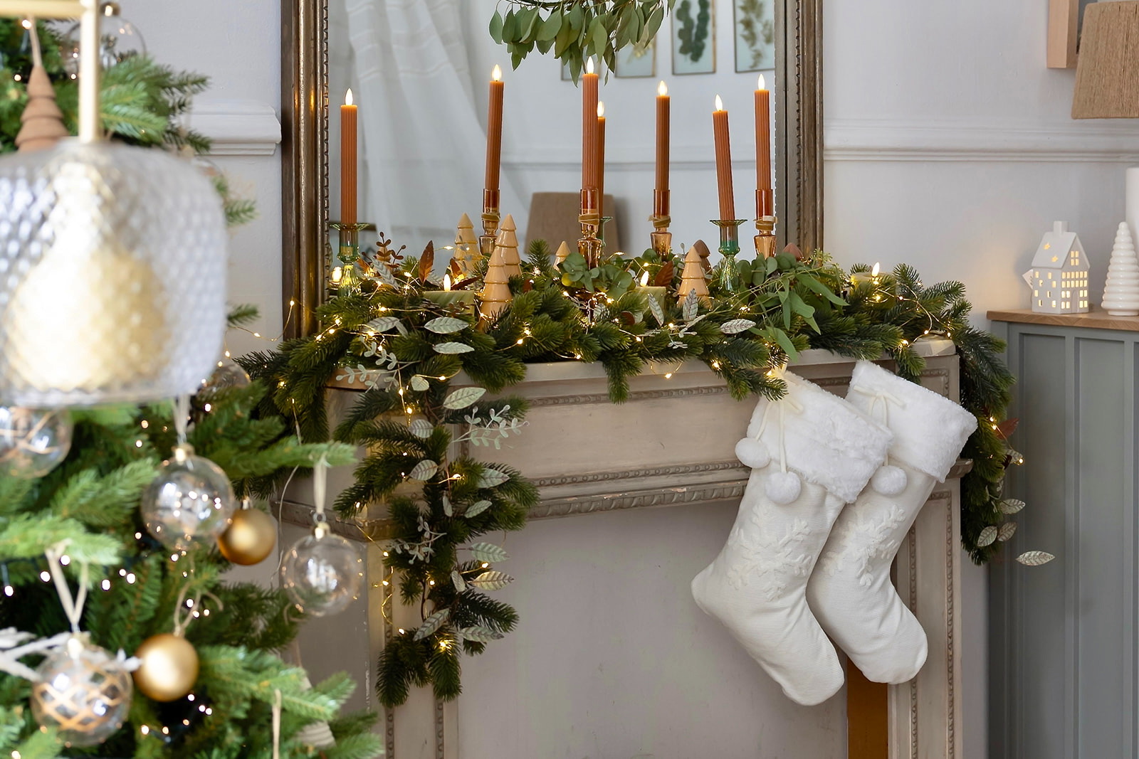 Decoración navideña elegante en chimenea con guirnalda de pino, velas doradas, calcetines blancos y luces brillantes. Incluye adornos dorados, pequeñas casas iluminadas y elementos naturales como hojas verdes.