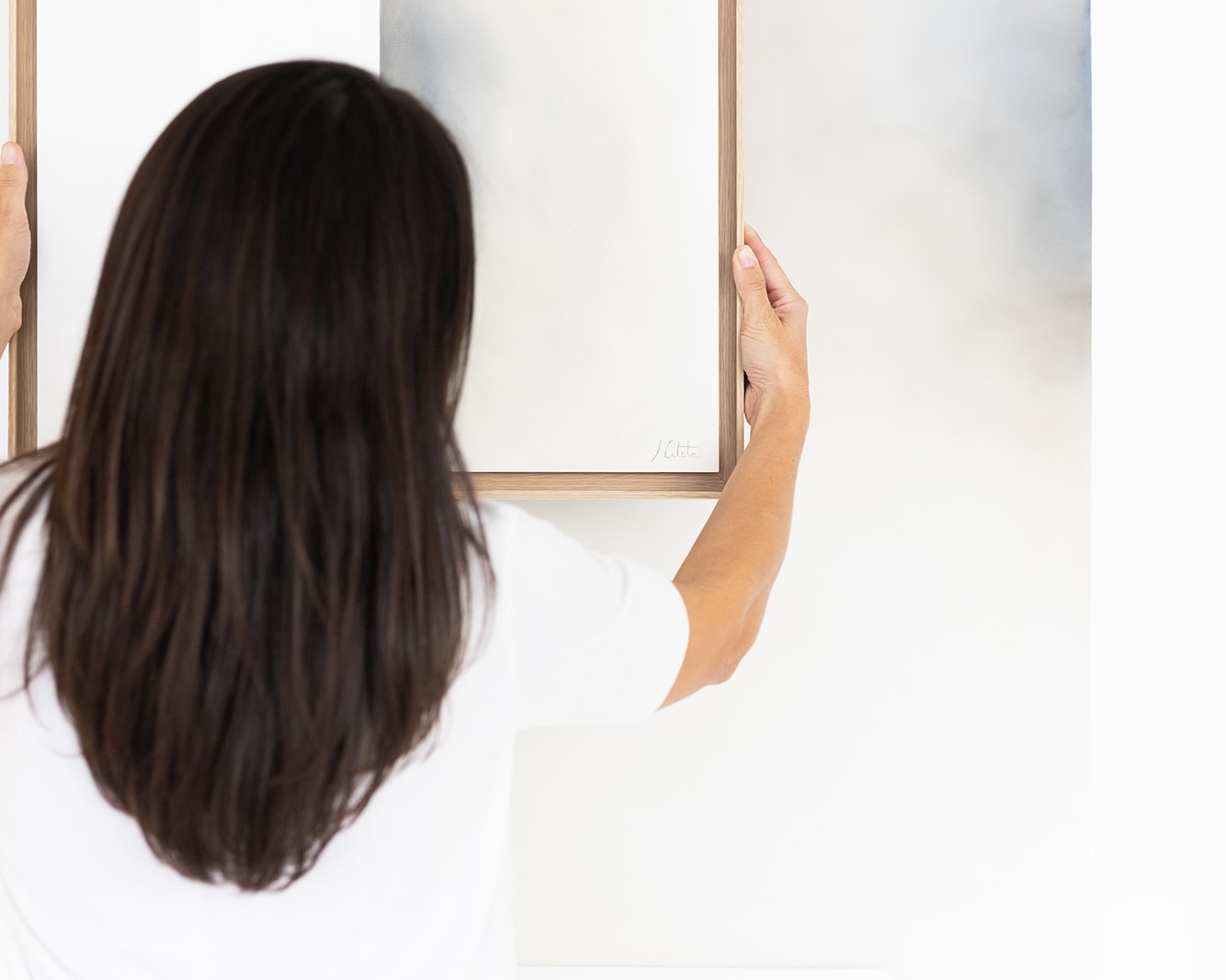 Vista trasera de una mujer de cabello largo y oscuro colgando un cuadro con marco de madera en una pared blanca. La mujer viste una camiseta blanca y sostiene el cuadro con ambas manos.