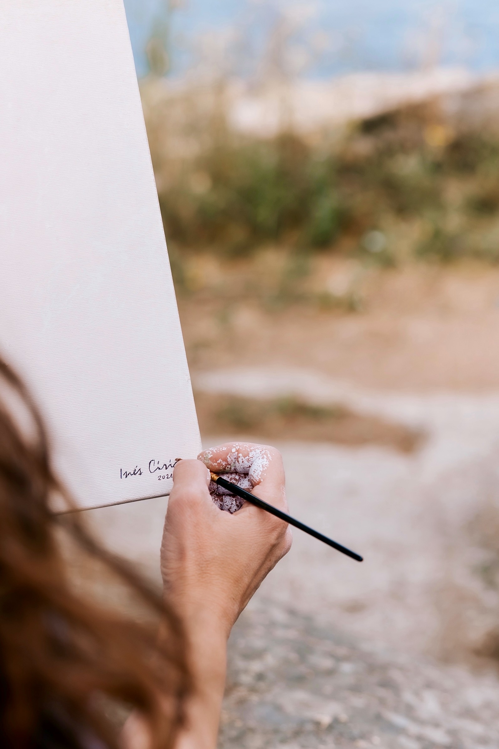 Una mano sosteniendo un pincel firma un lienzo blanco en un entorno de playa. El fondo muestra arena y vegetación costera borrosa.
