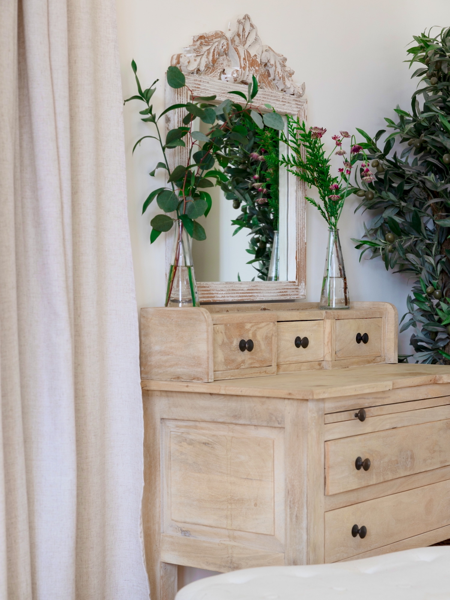 Cómoda vintage de madera clara con espejo decorativo y jarrones de cristal con plantas verdes y flores. El mueble tiene varios cajones con tiradores negros y está decorado con un estilo rústico elegante.