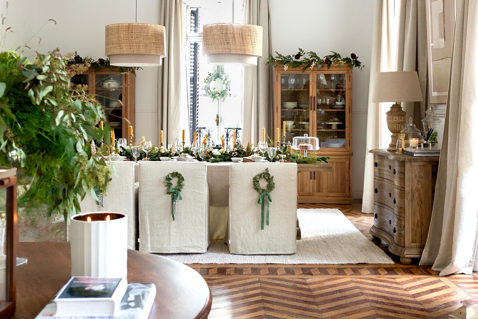 Comedor decorado para Navidad con estilo elegante y natural, mesa con fundas blancas, coronas verdes, velas doradas, lámparas colgantes con pantallas de yute y muebles de madera rústica sobre suelo de espiga