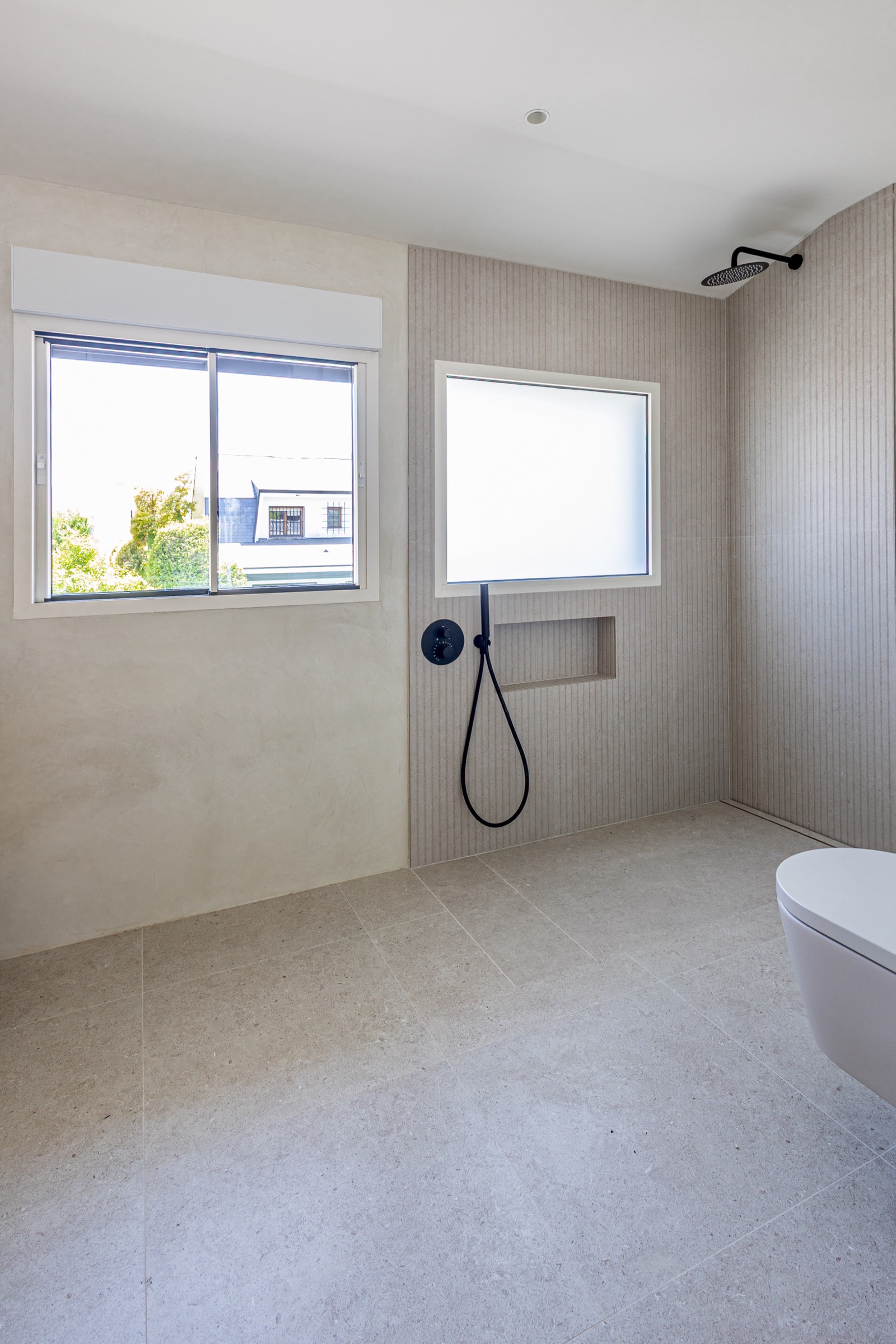 Cuarto de baño espacioso con paredes neutras, azulejos grises, ventana grande, ducha con cabezal negro y inodoro blanco flotante