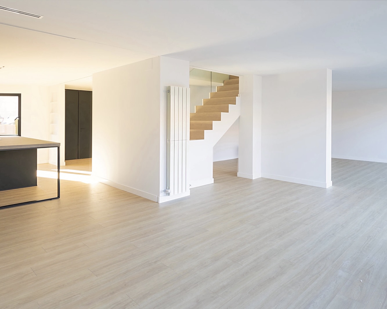 Interior minimalista con paredes blancas, piso de madera clara y una escalera de madera visible. El espacio es amplio y luminoso, con un diseño limpio y moderno.