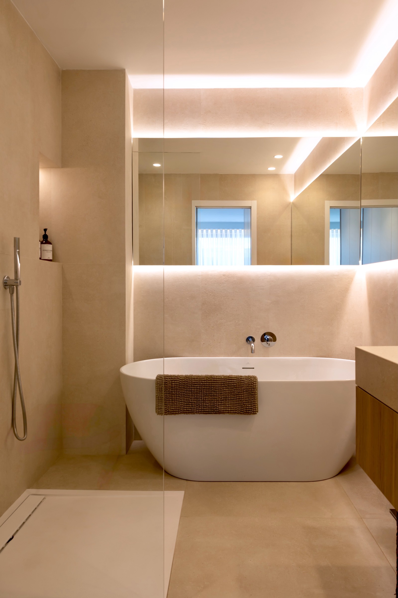 Baño moderno con bañera independiente blanca, paredes beige, iluminación indirecta en el techo, gran espejo y ventana que permite ver el exterior. Diseño minimalista y elegante con acabados de alta calidad.
