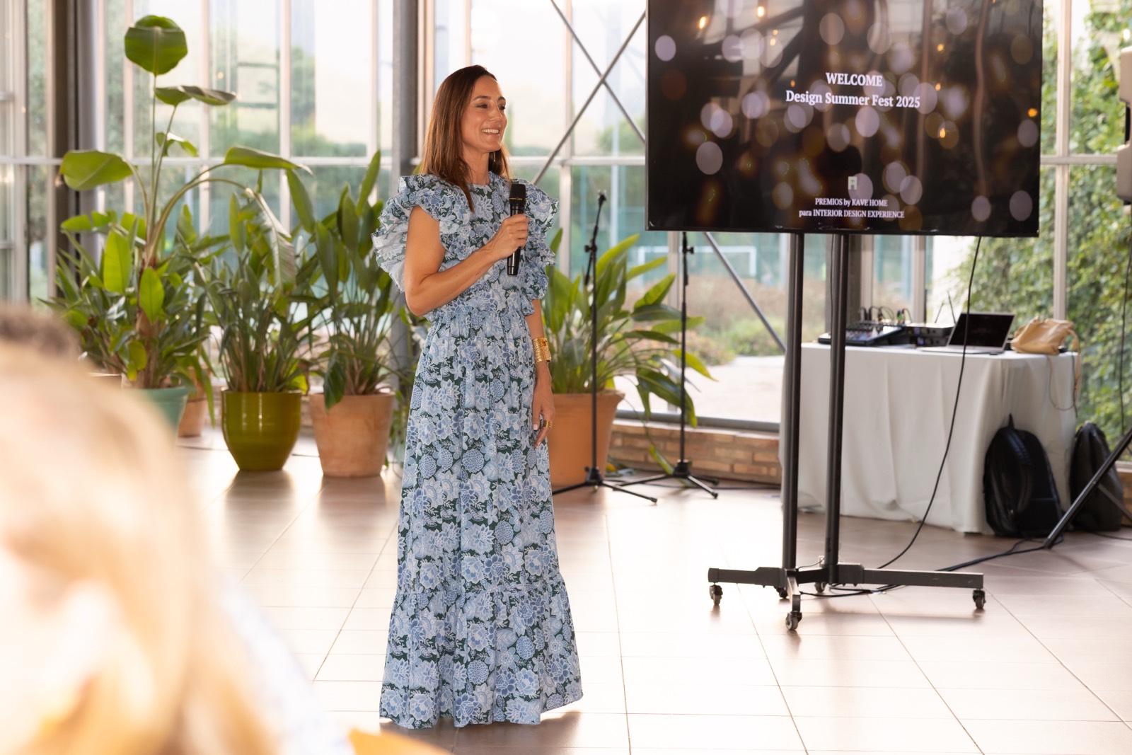 Presentadora con vestido floral azul hablando en un evento de Design Summer Fest 2025, con plantas verdes y una pantalla de bienvenida