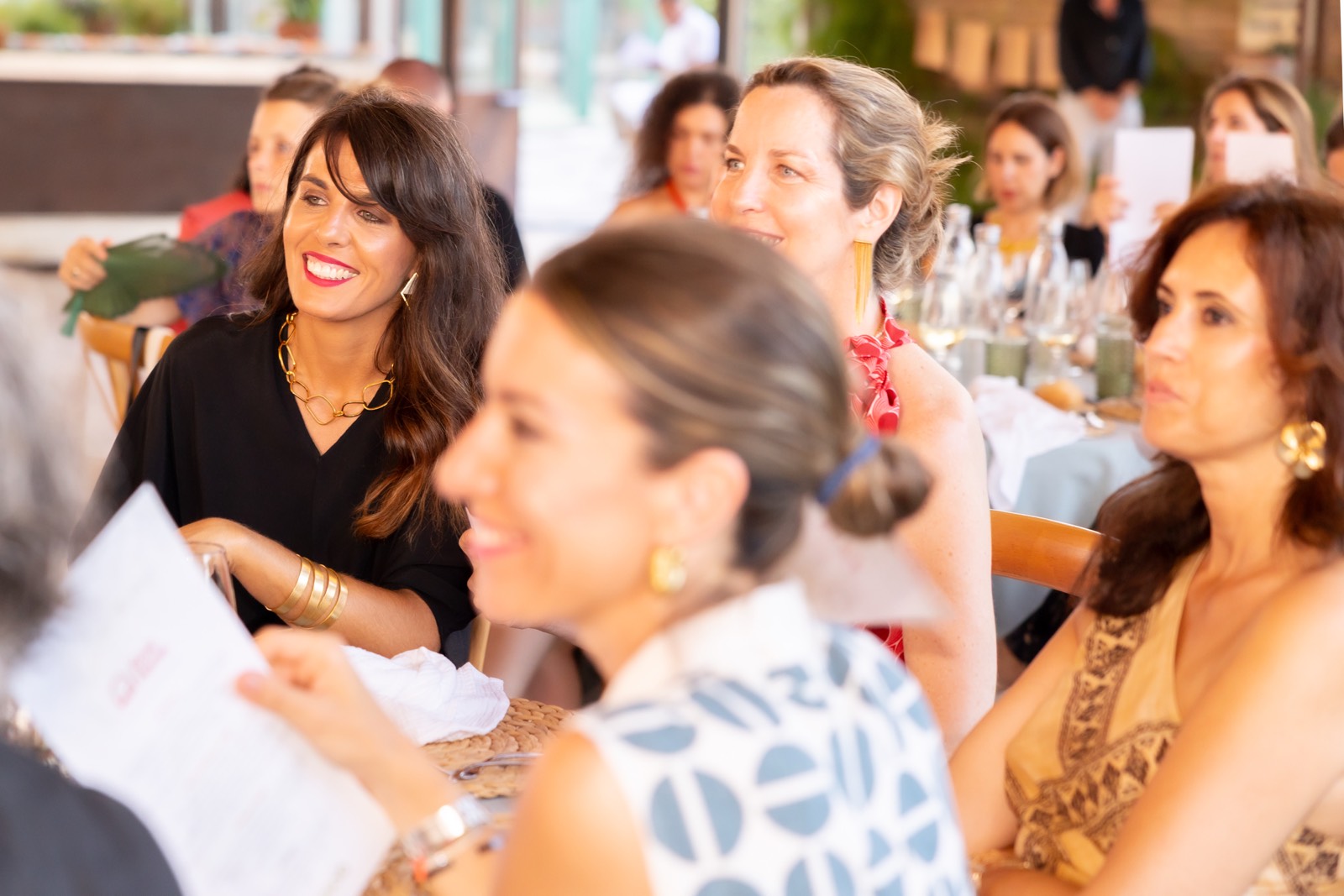 Grupo de mujeres reunidas en un evento, sonriendo y conversando alrededor de una mesa con botellas y vasos