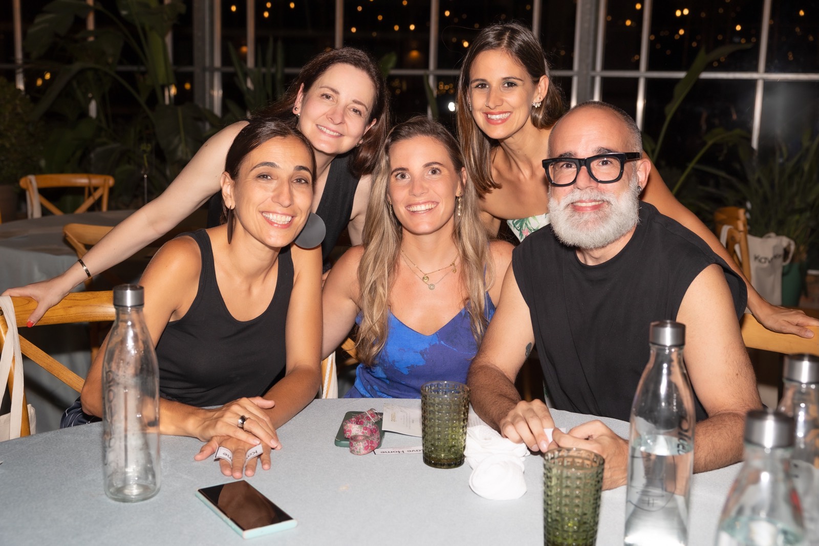 Grupo de cinco amigos sonrientes sentados juntos en una mesa con botellas de agua y vasos, en un ambiente iluminado con luces nocturnas