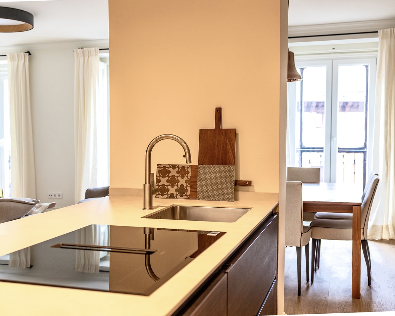 Cocina moderna de planta abierta con isla, encimera beige, fregadero empotrado y grifo de acero inoxidable. Se conecta con un comedor luminoso con mesa de madera y sillas tapizadas. Decoración minimalista con tablas de cortar y azulejos decorativos.