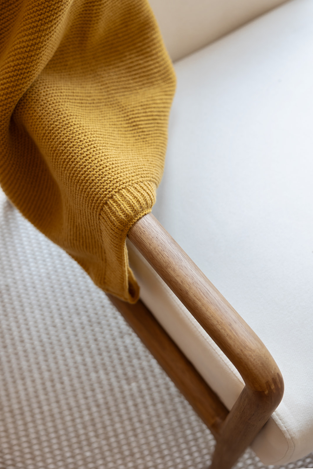 Detalle de una manta tejida de color mostaza sobre un sillón con estructura de madera. La textura del tejido es visible y contrasta con la suavidad de la madera del brazo del sillón.