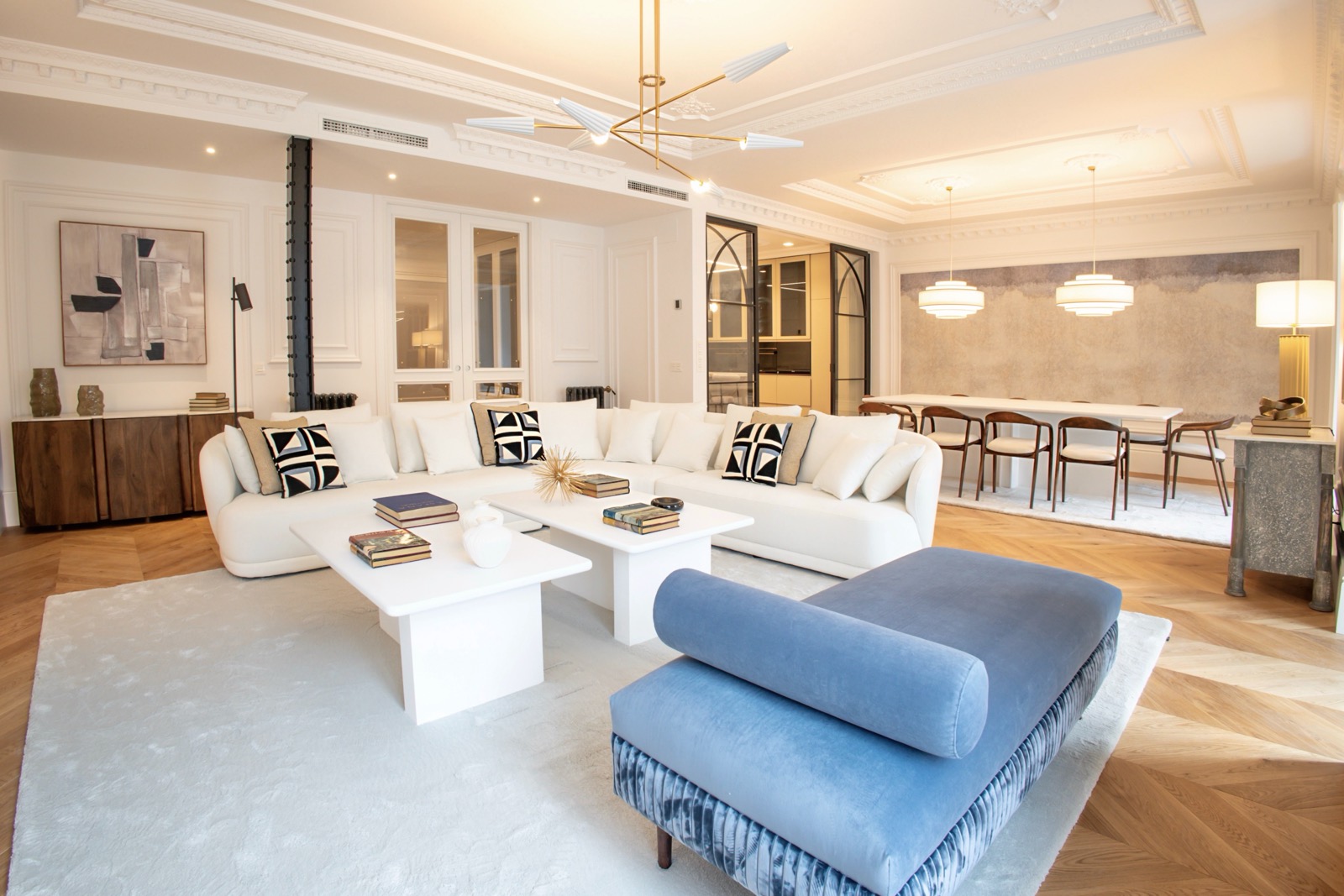 Amplia sala de estar con diseño moderno y elegante. Sofá blanco en L, mesas de centro blancas, otomana azul, lámparas colgantes y comedor al fondo. Decoración en tonos neutros con acentos en negro y azul.