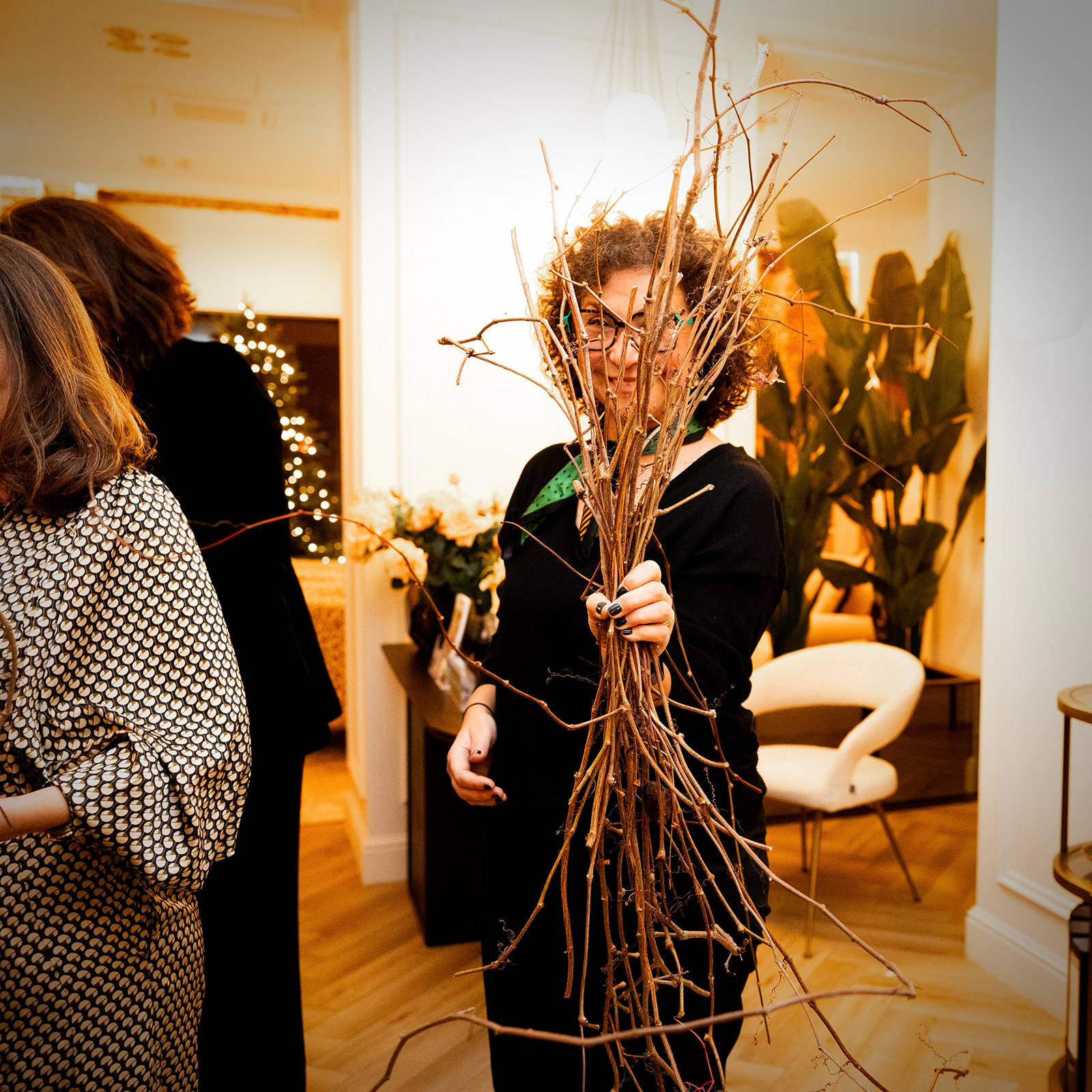 Persona sosteniendo un ramo de ramas secas en un evento interior. Ambiente cálido con decoración navideña y plantas. Otras personas presentes en el fondo.